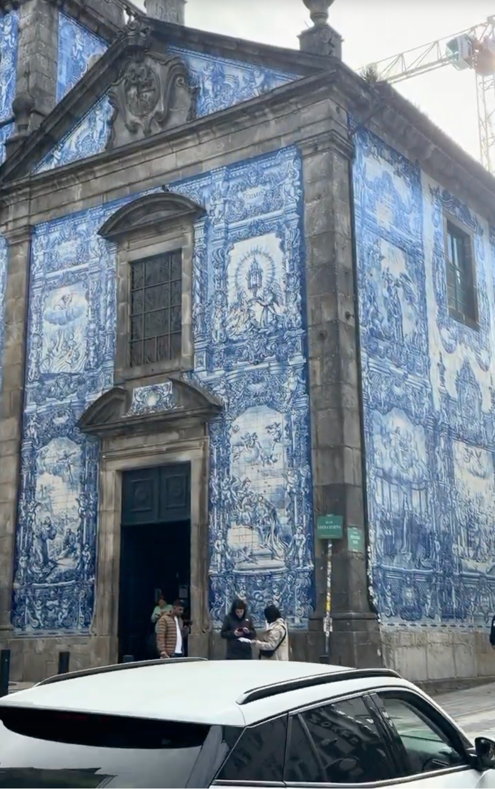 A church with blue and white tiled murals covering its walls, featuring intricate designs and religious scenes. A few people stand near the entrance, and a white car is parked in front of the building.