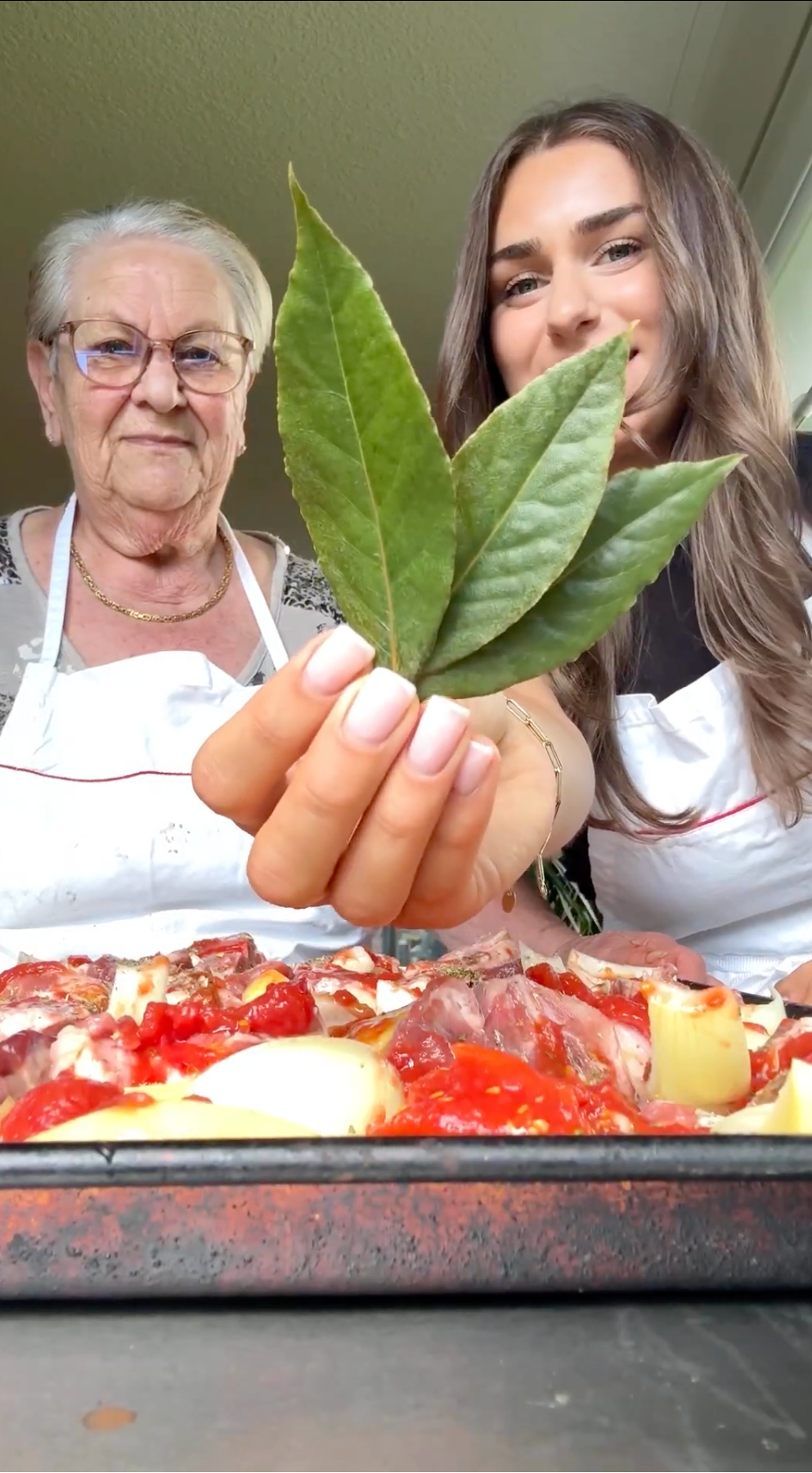 Two women in aprons stand behind a tray of food, smiling. One woman holds three large green bay leaves toward the camera, partially covering their faces. The scene appears to be in a kitchen.