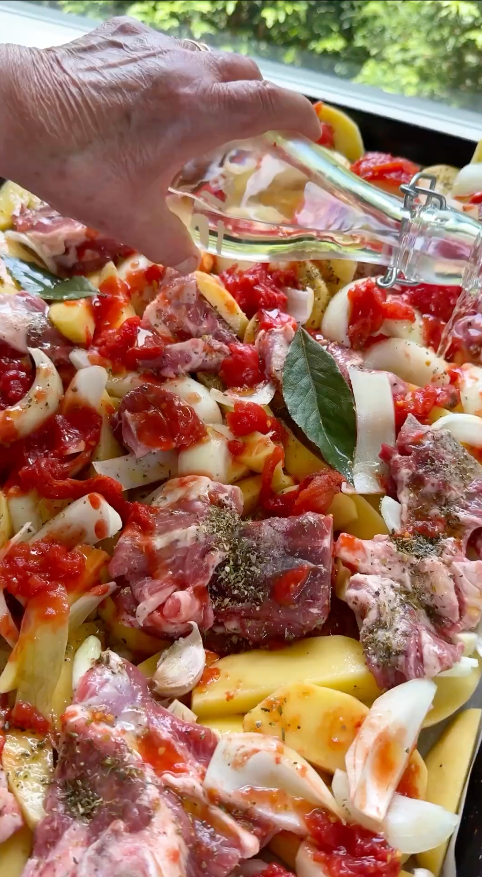 Chopped raw meat, potatoes, onions, bay leaves, tomatoes, and herbs are arranged on a tray. A hand pours olive oil over the ingredients, preparing them for roasting. Greenery is visible through a window behind the tray.