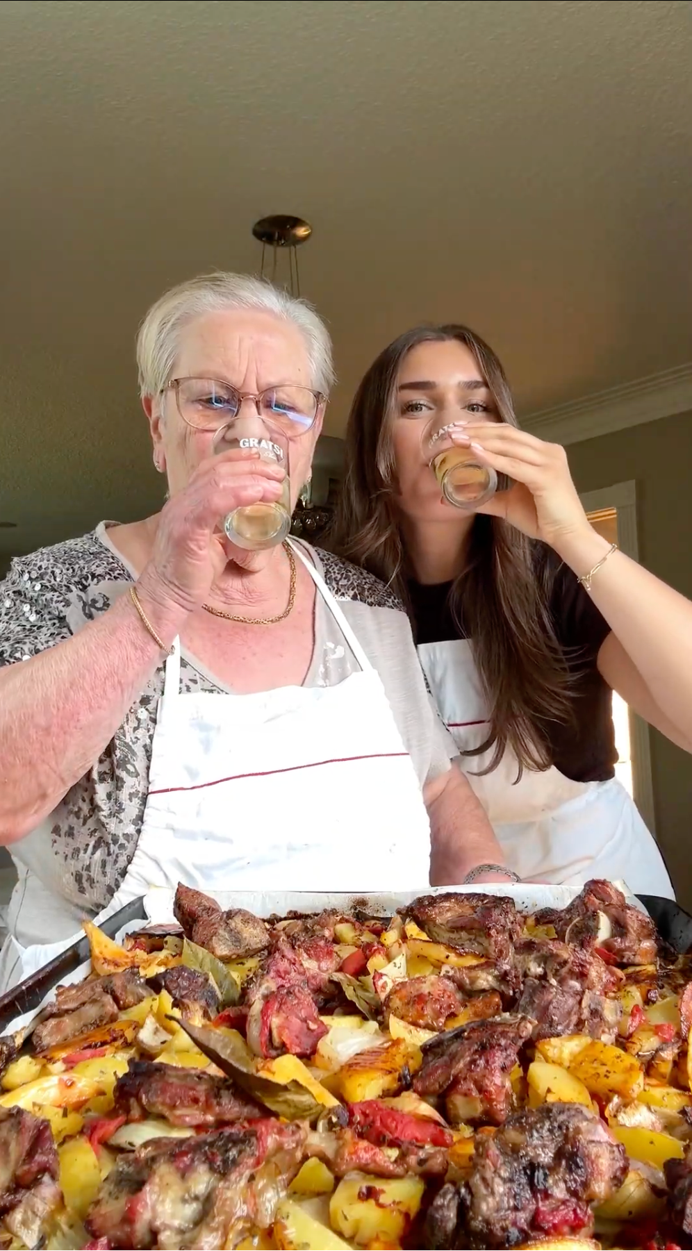 Two women, one older and one younger, wearing aprons and holding drinks, stand behind a tray of roasted meat and vegetables in a kitchen, smiling and enjoying their time together.