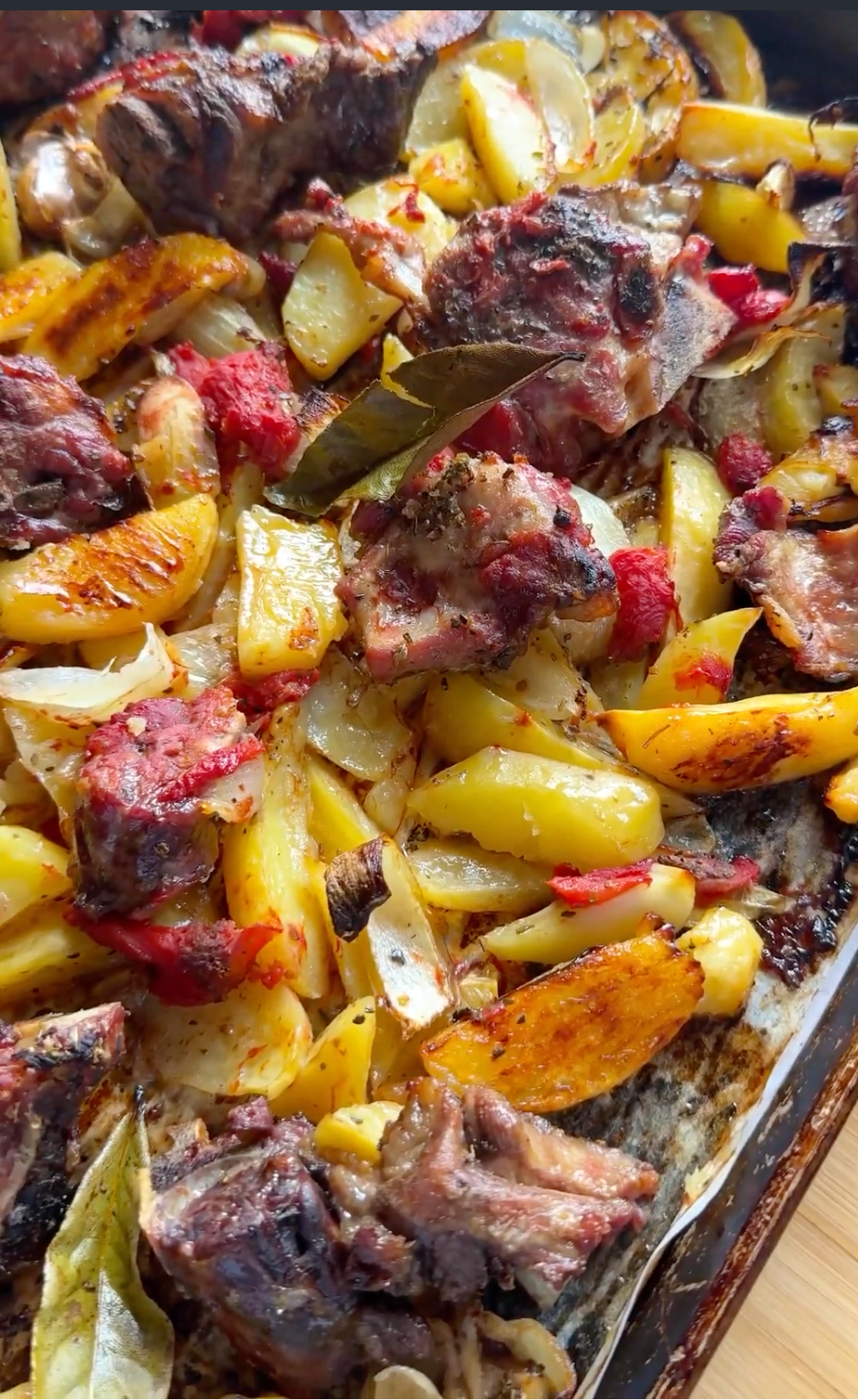 A close-up of a baked dish with roasted potatoes, onions, chunks of meat, tomato pieces, and bay leaves on a metal tray, showing a mix of golden, red, and brown colors.