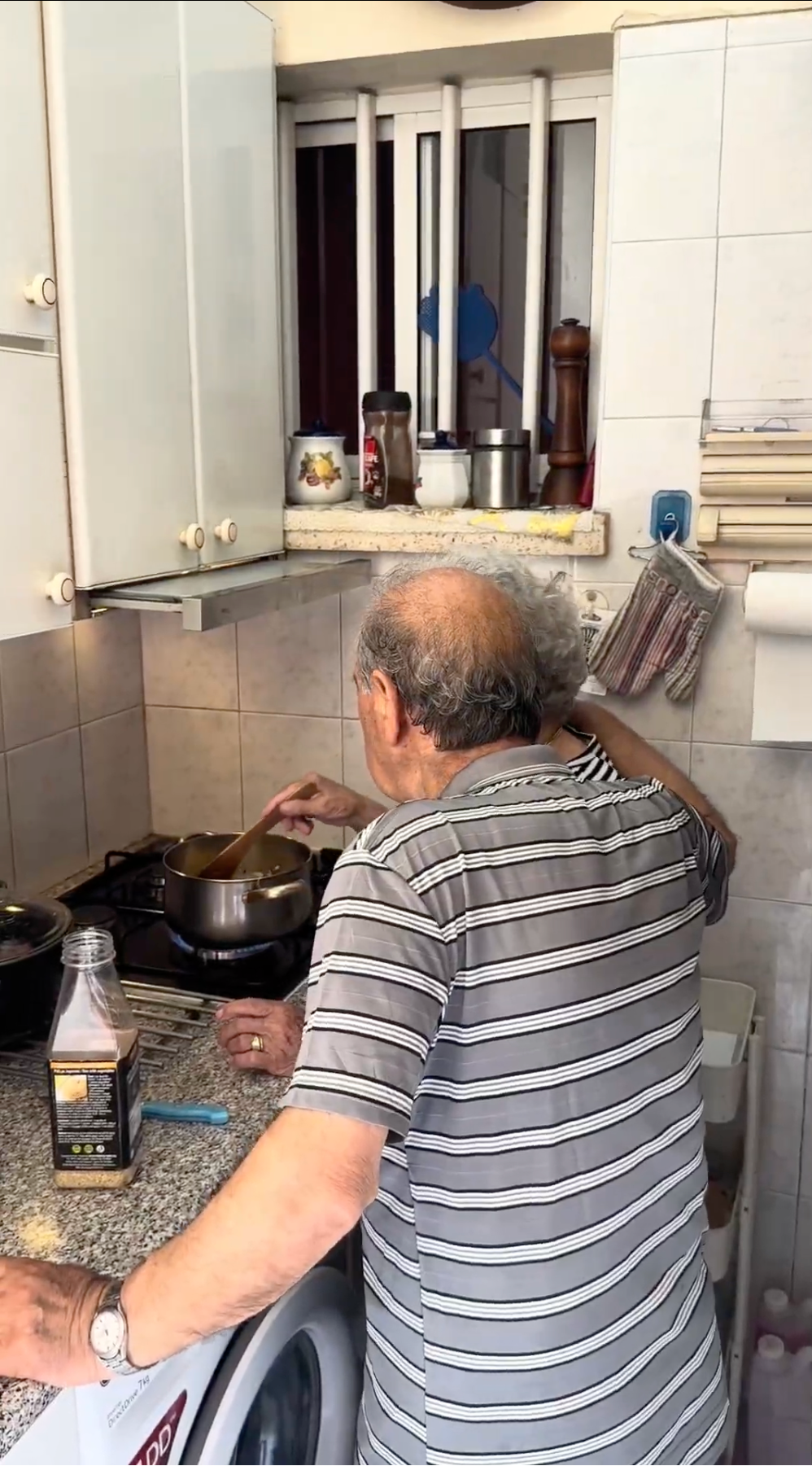 An elderly man with gray hair, wearing a striped shirt, cooks at a small kitchen stove, stirring a pot. Various kitchen items and condiments are on the counter and shelves around him.