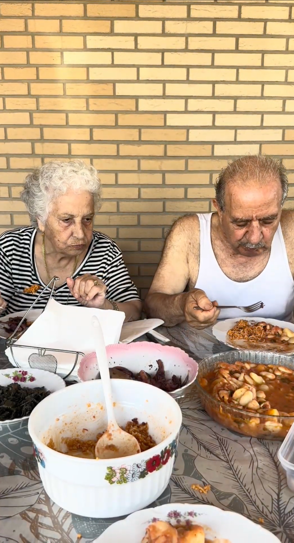 An elderly woman and man sit at a table with various dishes, eating a meal together. The table is covered with bowls and plates of food, and a brick wall is in the background.