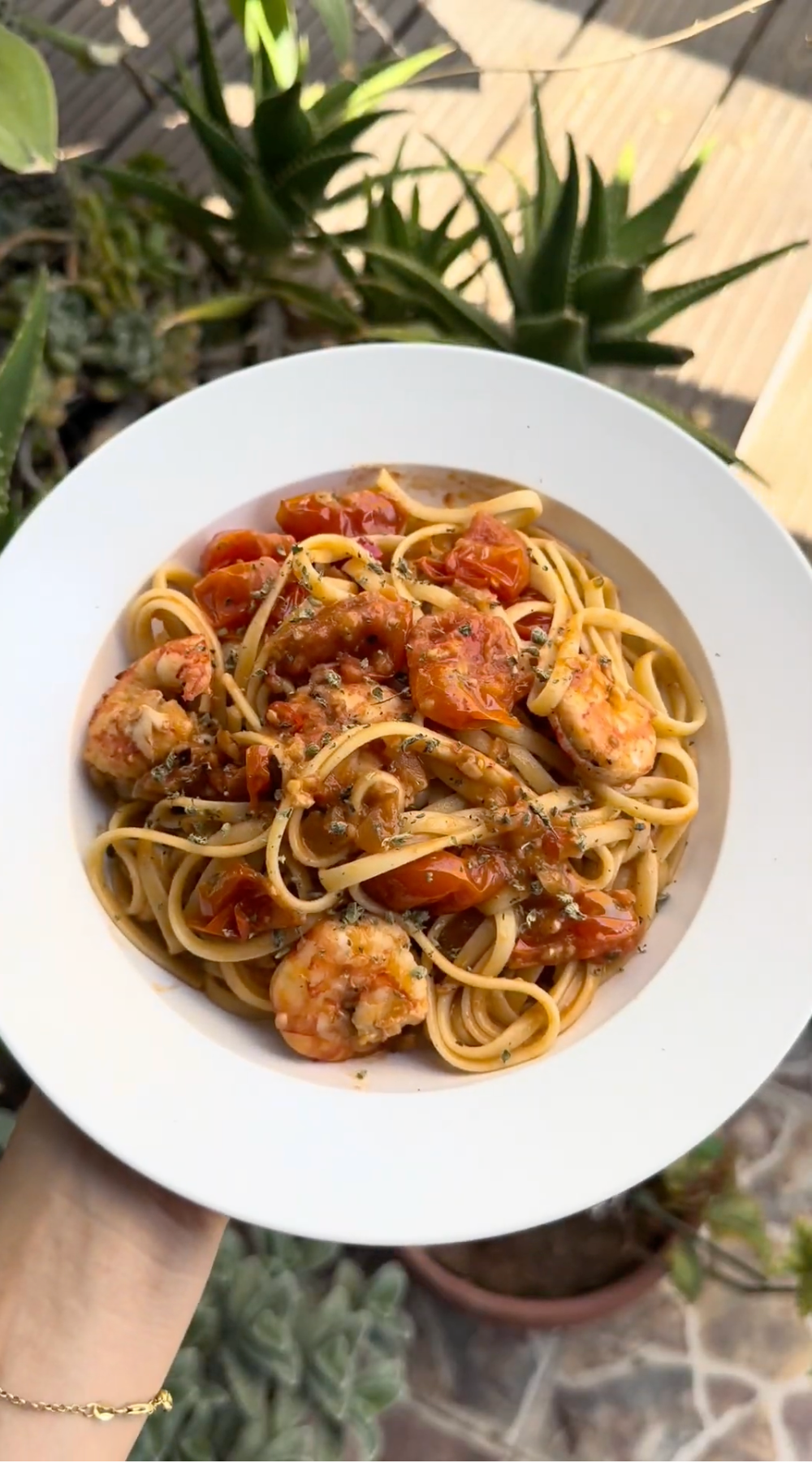 A white bowl filled with linguine pasta topped with shrimp, cherry tomatoes, and herbs is being held above a background of green plants.