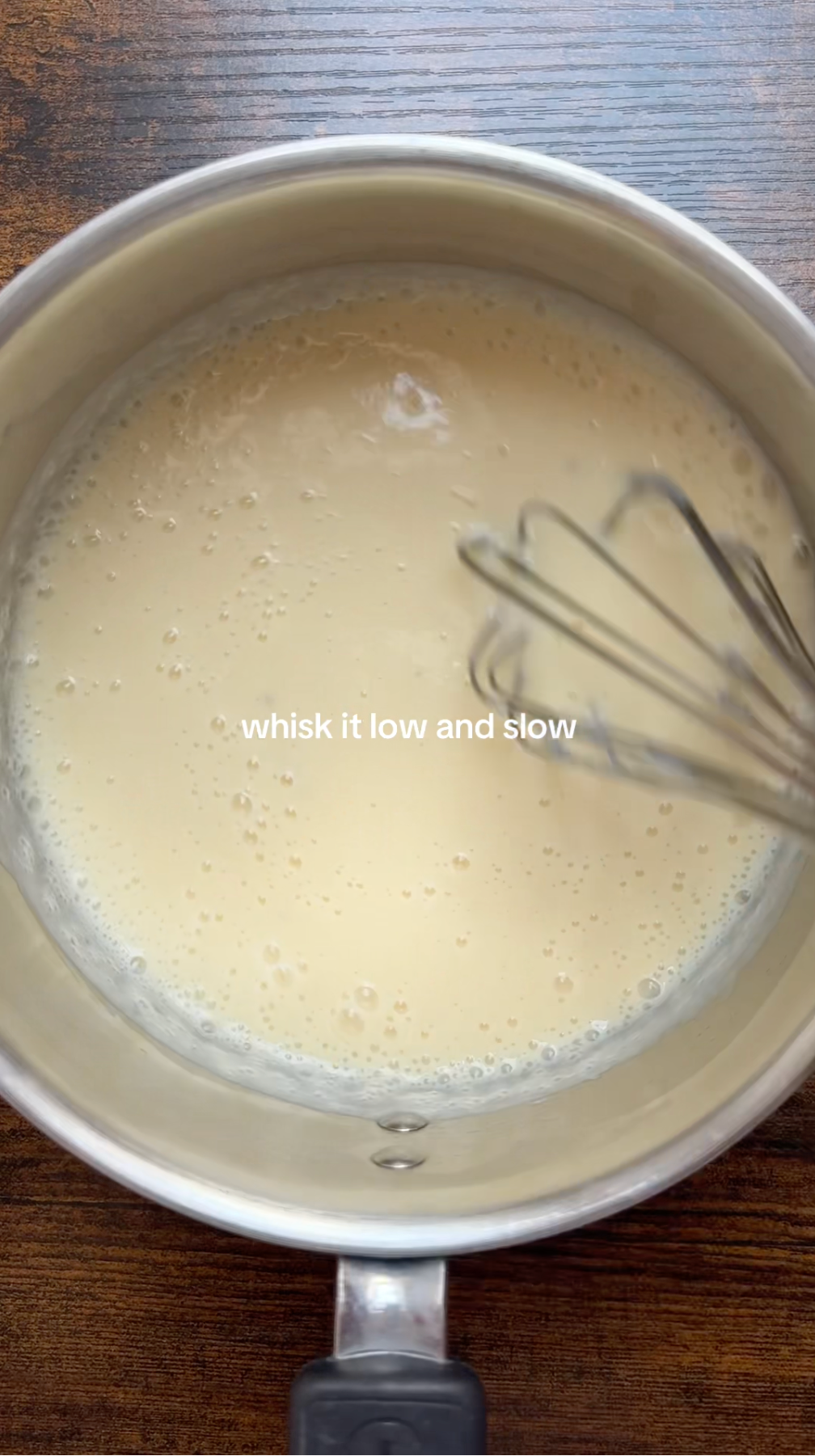 A metal whisk stirs a creamy light-yellow mixture in a stainless steel pot on a wooden surface. Text in the center reads, whisk it low and slow.