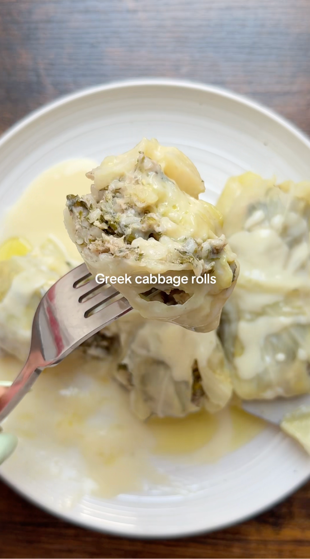 A fork holds a Greek cabbage roll topped with creamy sauce over a white plate with more cabbage rolls, all covered in sauce. Text on the image reads “Greek cabbage rolls.”.