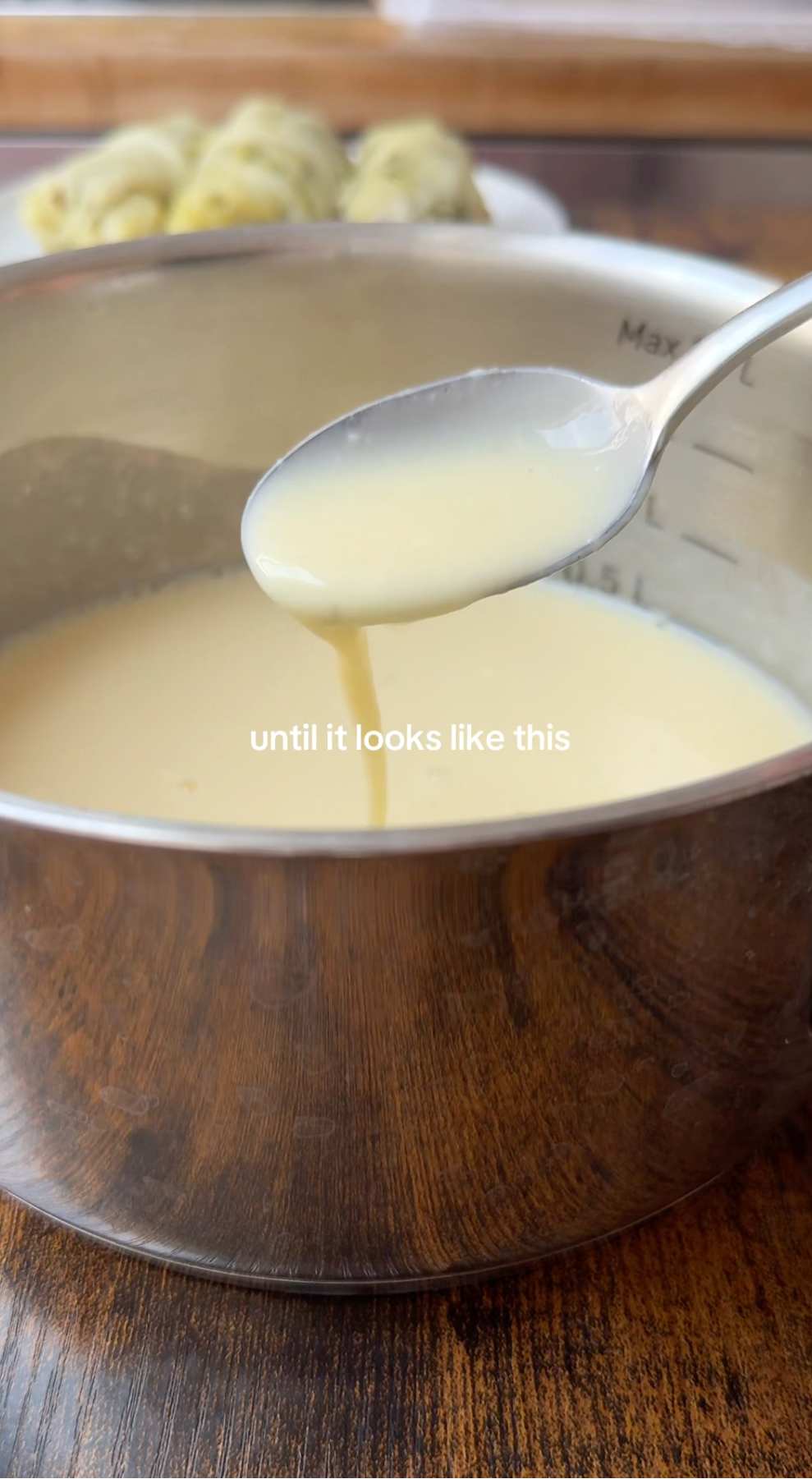 A spoon lifts creamy, pale yellow sauce from a metal saucepan on a wooden table. White text over the sauce reads, until it looks like this. Blurred food is visible in the background.