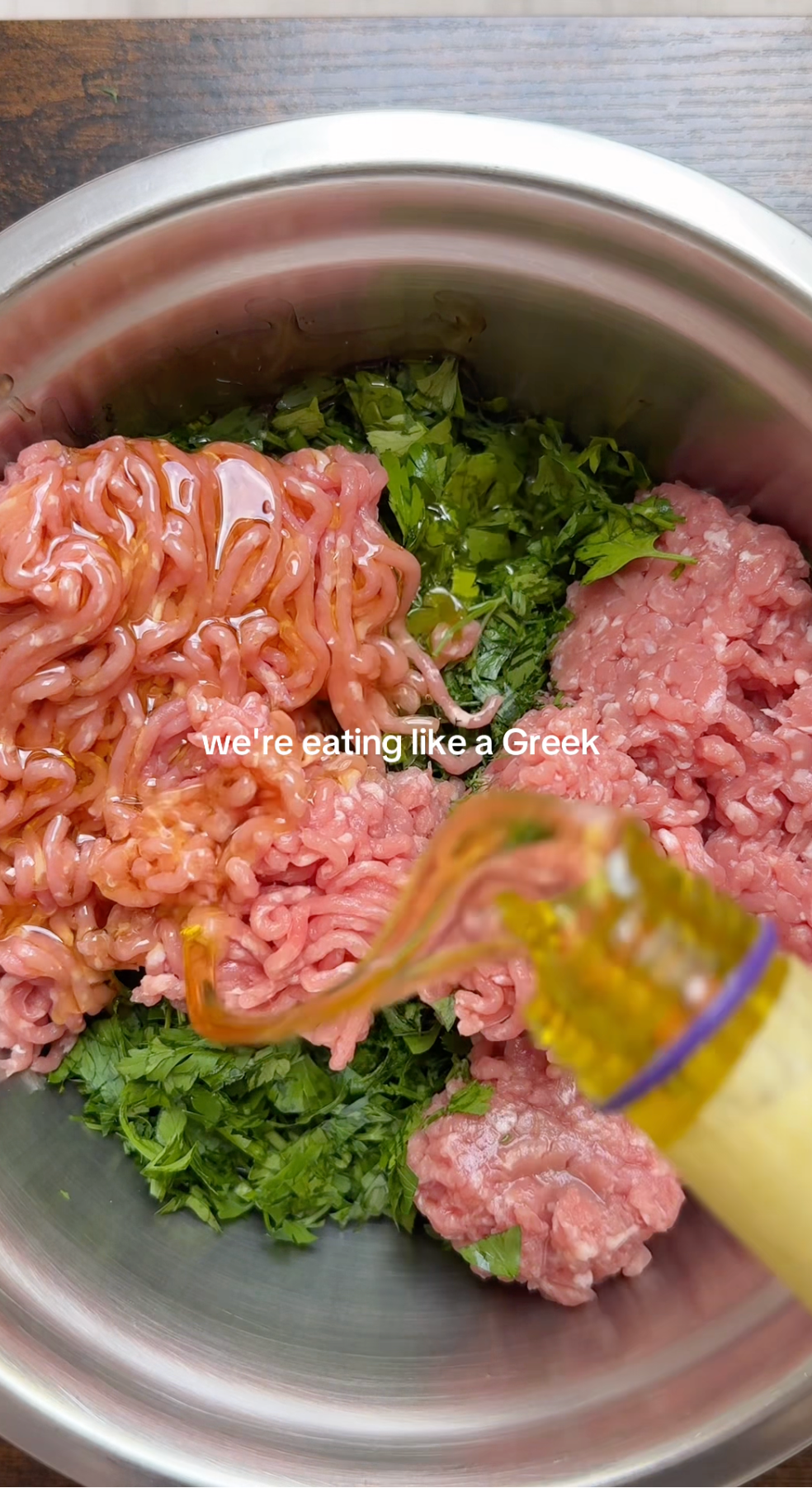 Ground meat, chopped parsley, and oil being poured into a metal bowl, with text overlay reading were eating like a Greek.