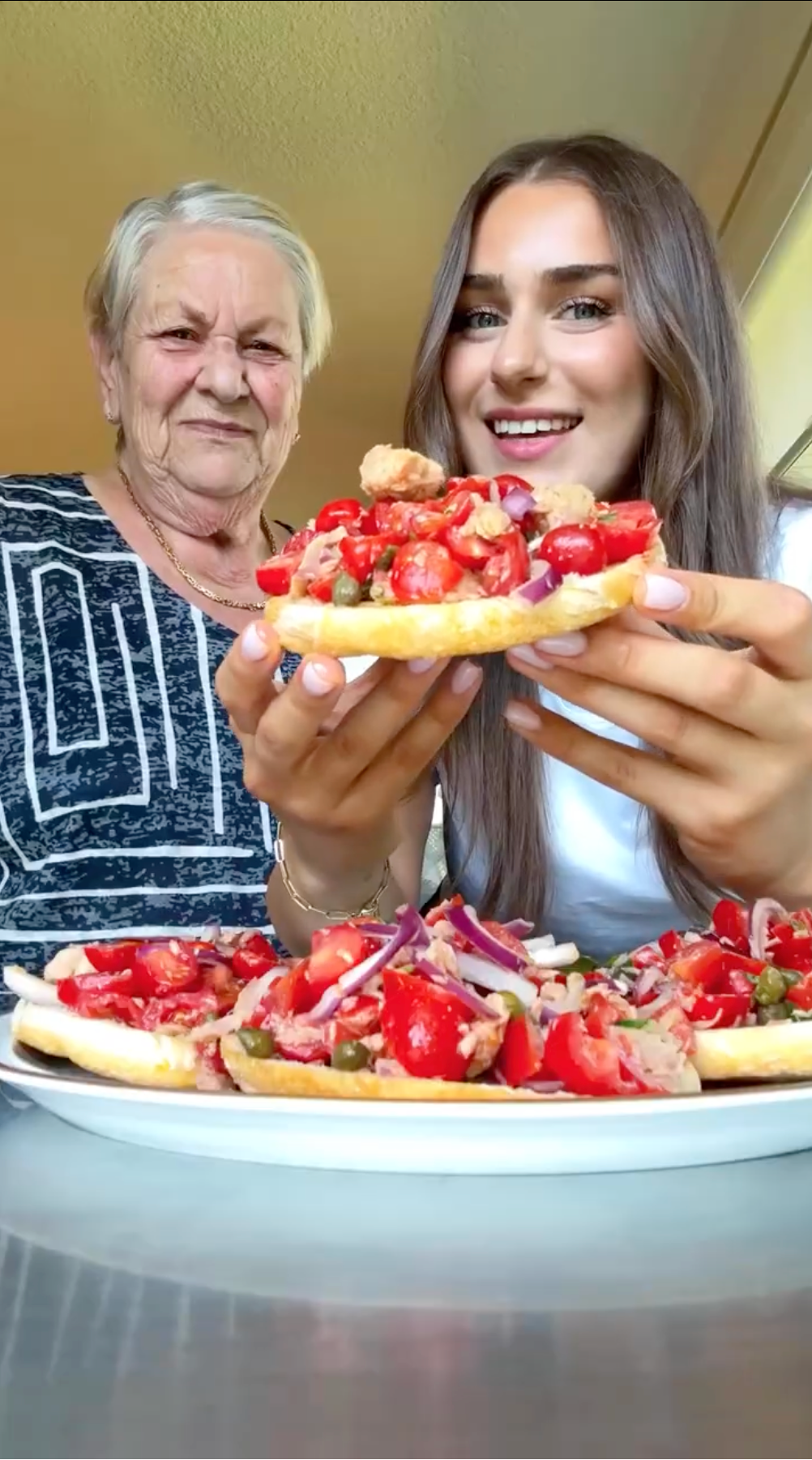 An older woman and a young woman smile at the camera. The young woman holds a slice of bread topped with tomatoes, onions, and meat. More topped bread slices are on the plate in front of them.