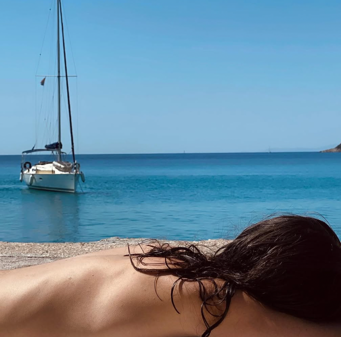 A person with wet dark hair lies on a stone surface by a calm blue sea, with a sailboat anchored nearby and a clear, sunny sky overhead.