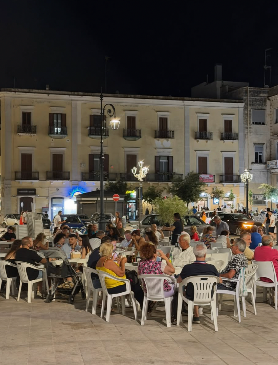 People sit at outdoor tables on a well-lit plaza at night, surrounded by multi-story buildings with balconies and shops. Cars are parked nearby under streetlights. The atmosphere is lively and social.
