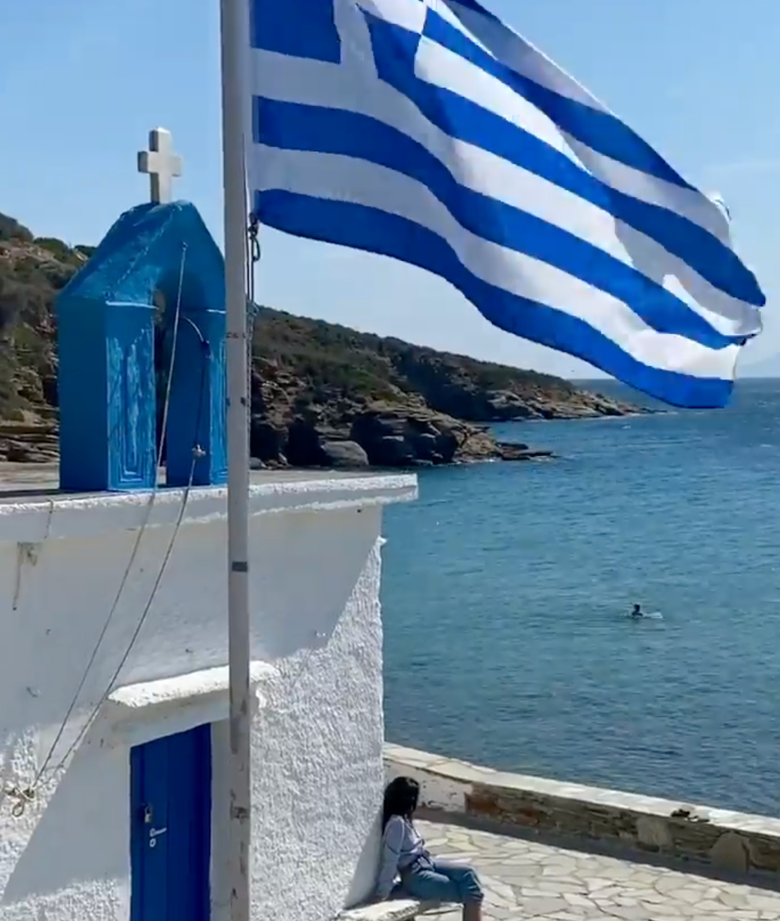 A Greek flag waves by a white chapel with a blue door and cross near the sea. A person sits on the chapel’s ledge, enjoying the coastal view under a clear sky.