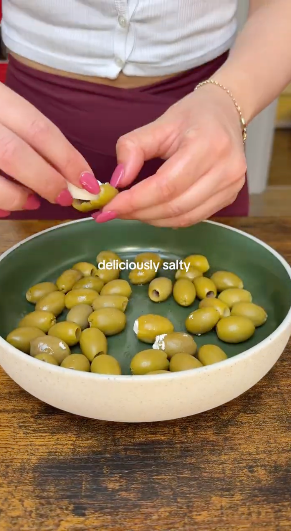 A person stuffs green olives with cheese over a bowl full of olives. Text on the image reads “deliciously salty.”.