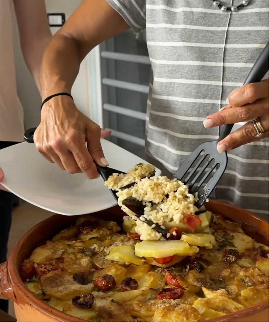A person serves a portion of baked rice and vegetables from a casserole dish onto a white plate, using a black spatula. Another person holds the plate.