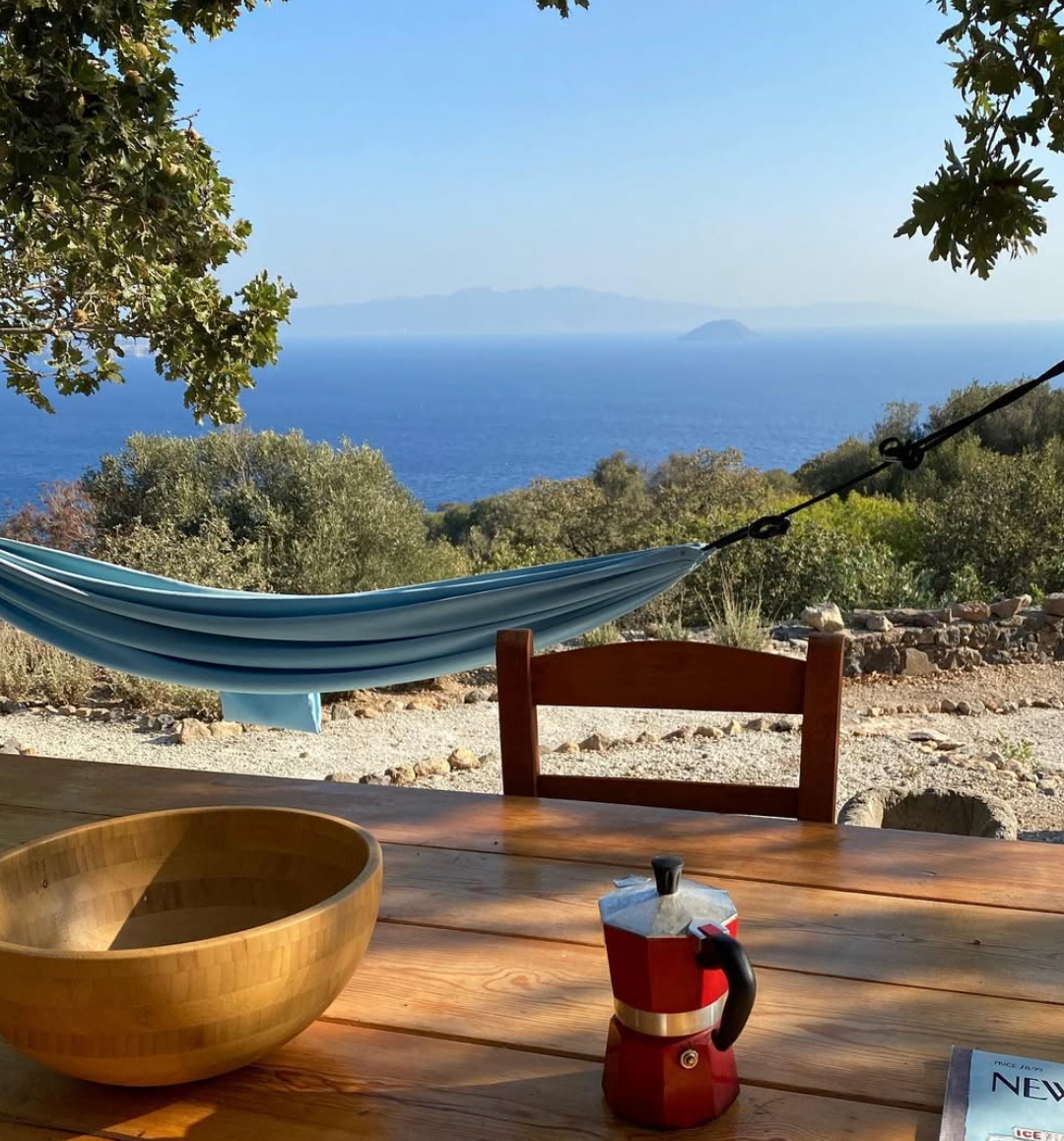 A wooden table with a bowl and red coffee pot overlooks a blue hammock, trees, and a sea view with distant mountains under a clear sky.