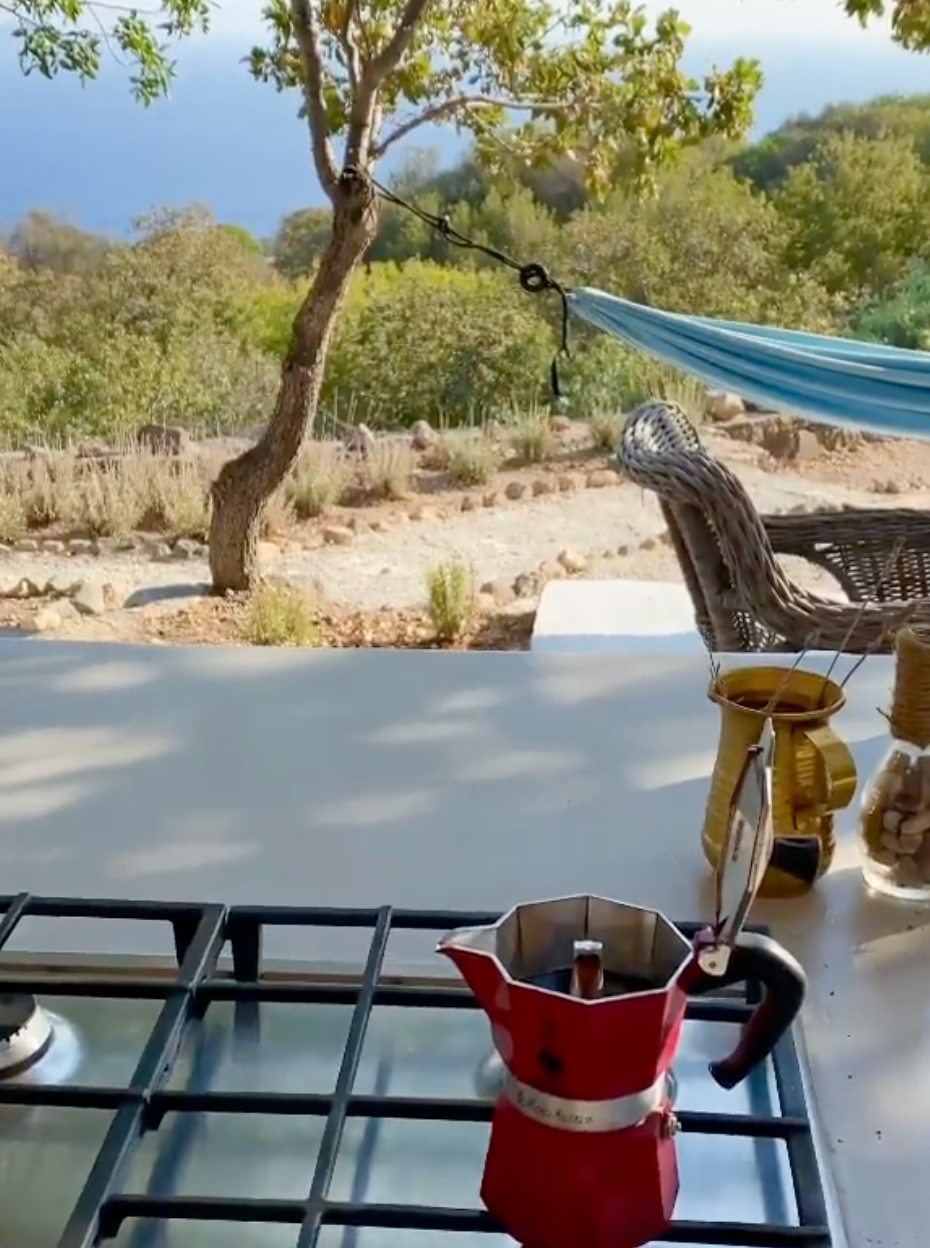 A red moka pot sits on a stove outdoors, with a hammock, wicker chair, and jars nearby. In the background are trees, rocky ground, and a glimpse of the sea under a clear sky.