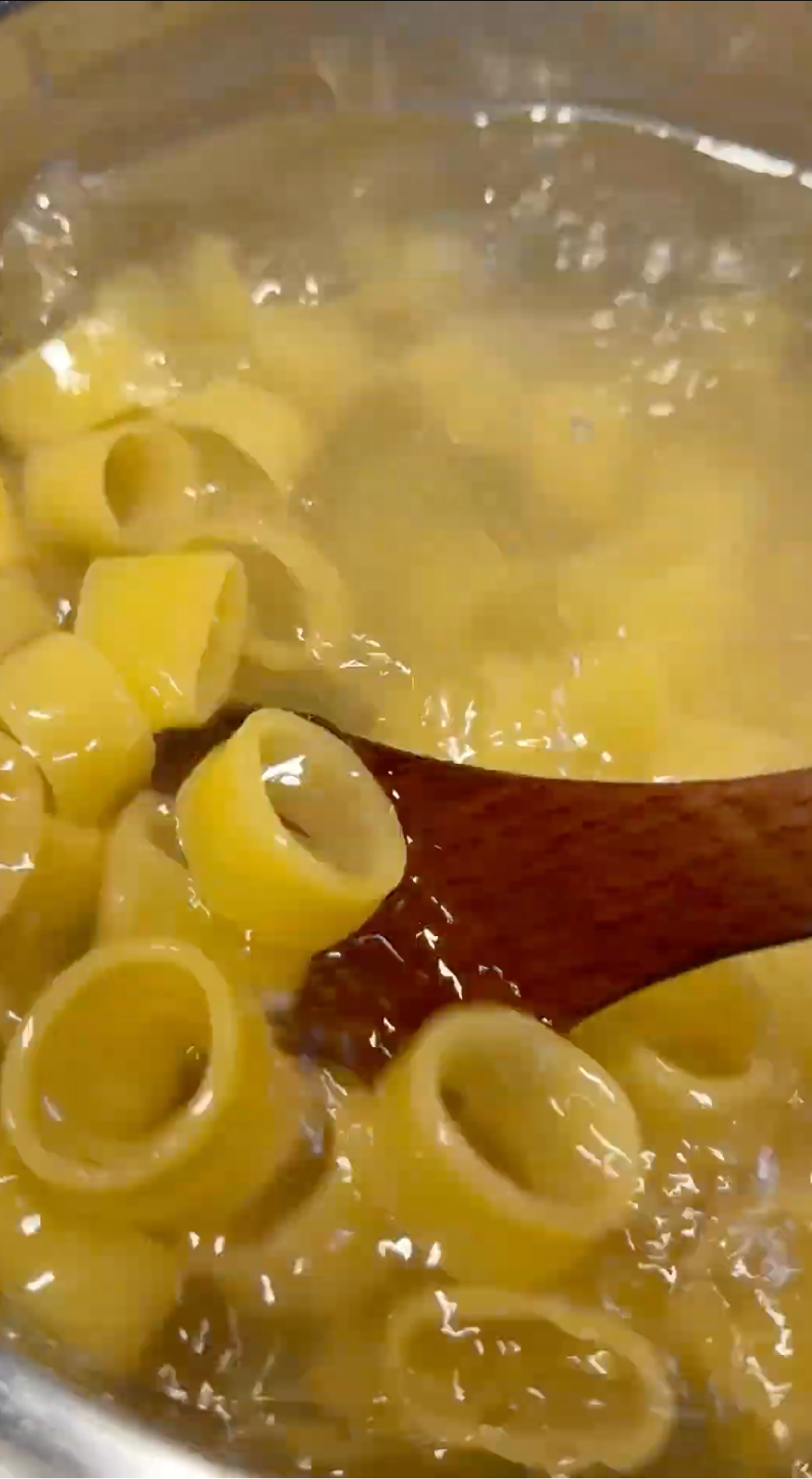 Large tube-shaped pasta is boiling in a pot of water, with a wooden spoon stirring the pasta. Steam rises from the pot.