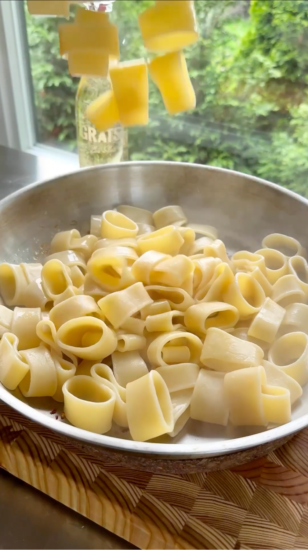 Large pasta noodles tumble down into a steaming pan of cooked pasta set on a wooden board, with a window and greenery visible in the background.