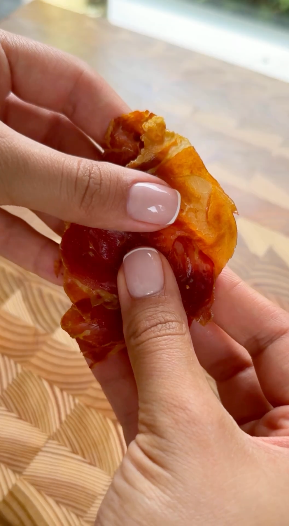 A person with neatly manicured nails holds and pulls apart a small pastry filled with red jam over a wooden patterned surface.