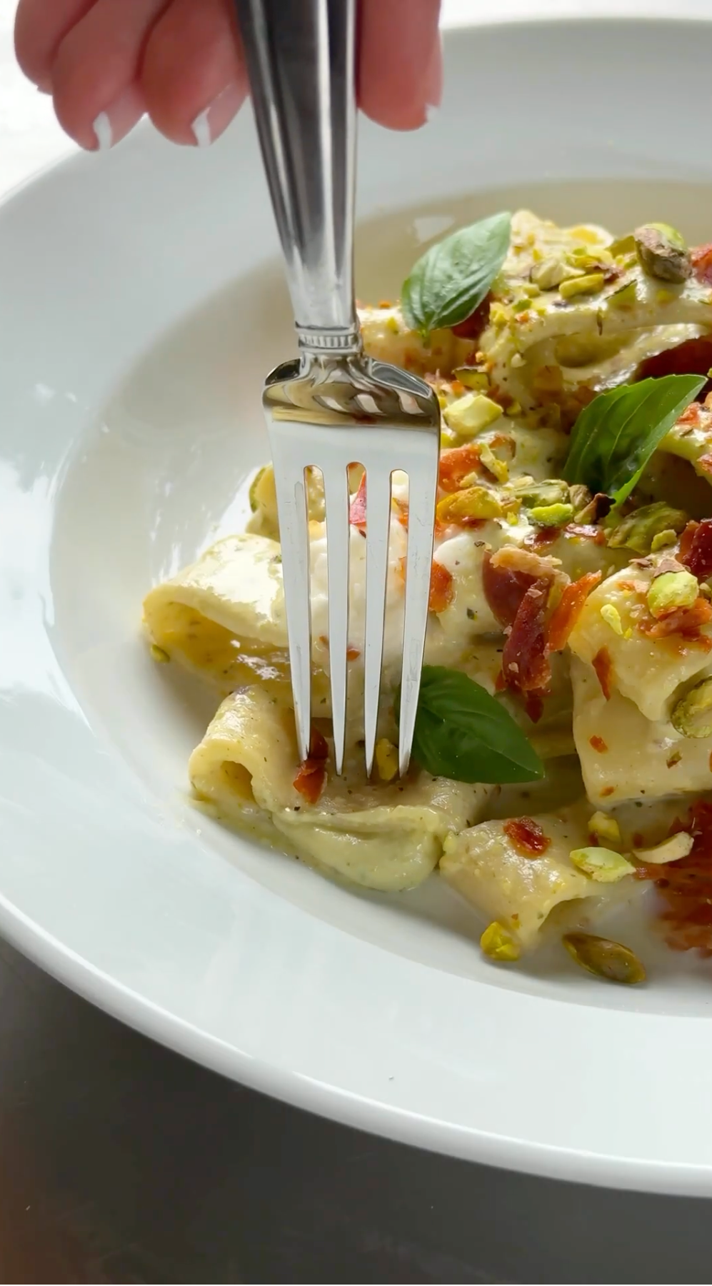 A hand holds a fork poised above creamy pasta garnished with basil, pistachios, and crispy bits, served in a white bowl.