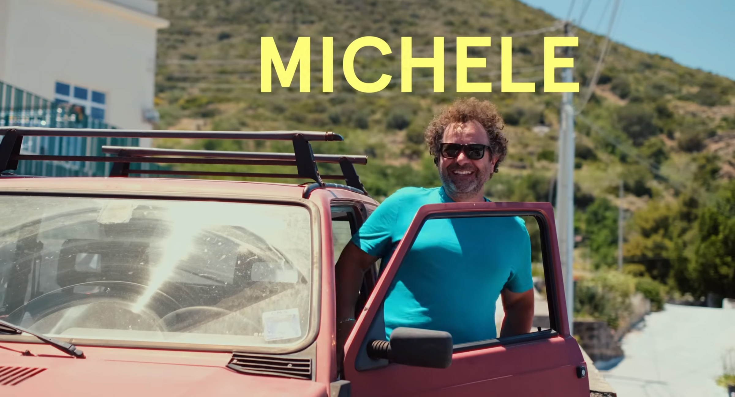 A man in sunglasses and a blue shirt smiles while standing by an open car door. The background shows a sunny hillside, and large yellow text above reads MICHELE..