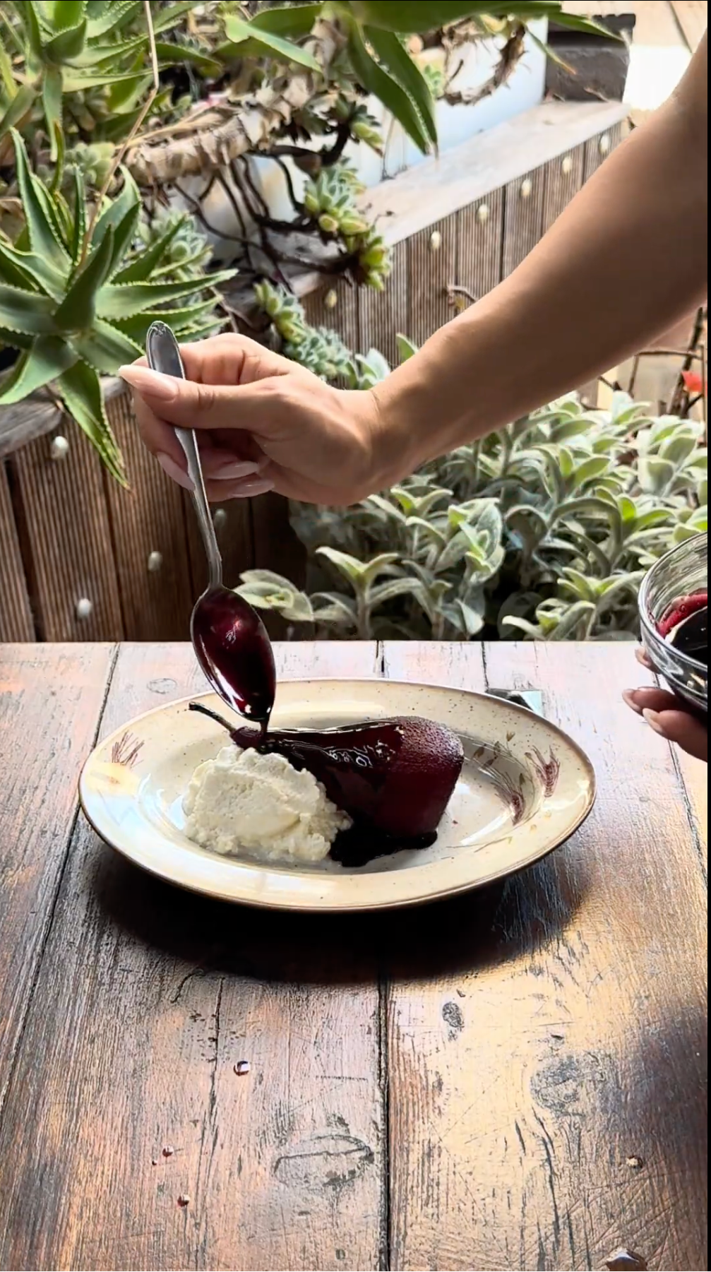 A hand spoons dark sauce over poached pear and whipped cream on a plate atop a rustic wooden table, surrounded by lush green potted plants.
