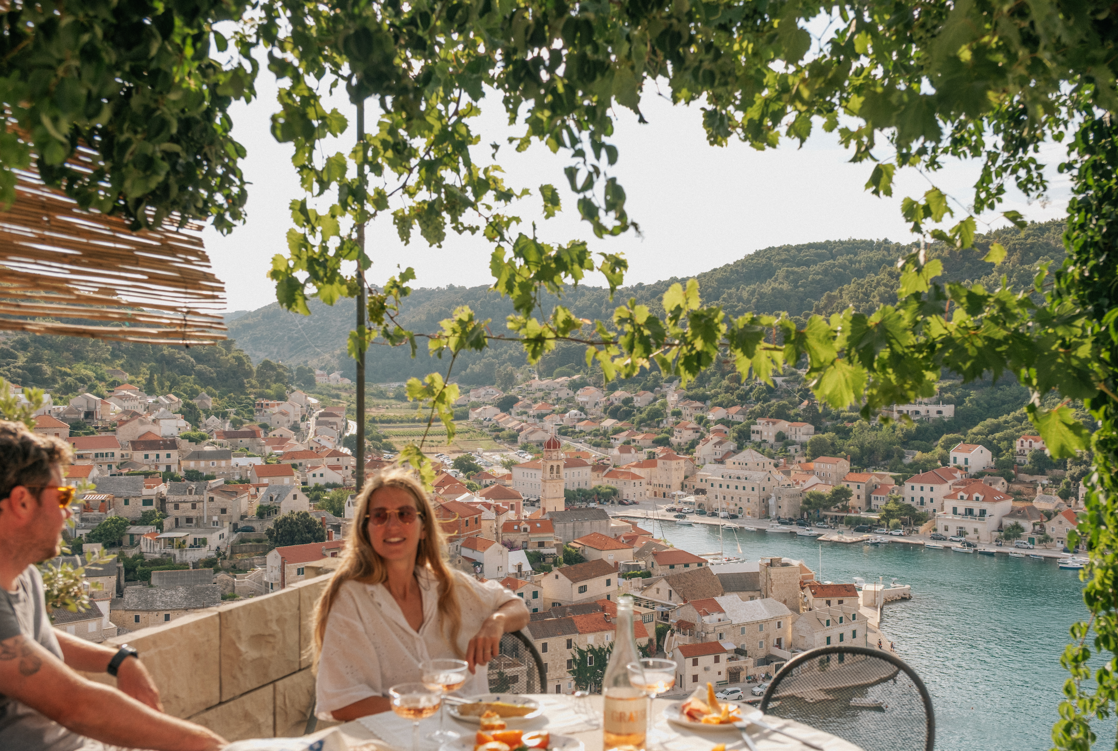 Two people sit at a terrace table with food and drinks, overlooking a scenic coastal village with red-roofed houses, hills, and water. Green vines hang above, partially shading the area.