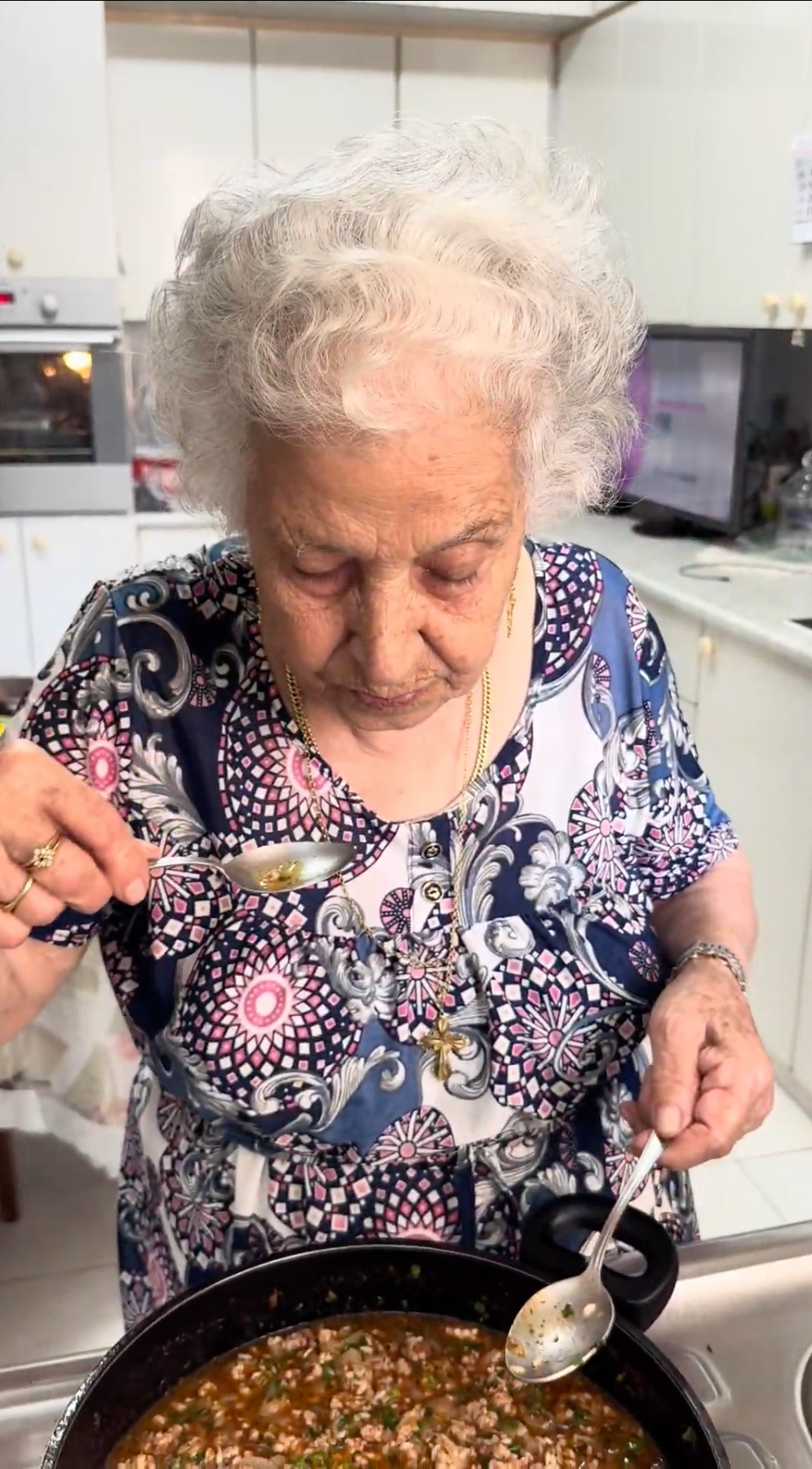 Elderly woman with white hair and a patterned blouse tastes food from a spoon in a kitchen, standing over a pot of stew.