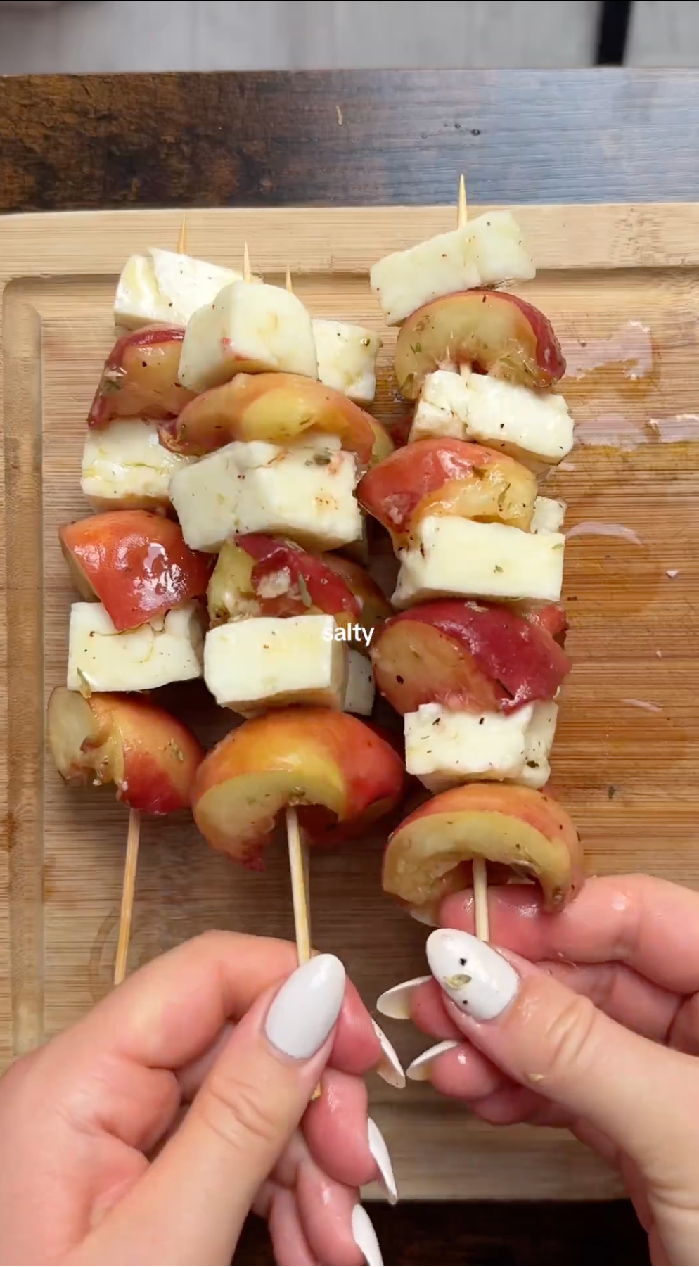 Two hands hold wooden skewers with alternating slices of white cheese and red apples, placed on a wooden cutting board. The food appears seasoned, and the skewers are neatly arranged side by side.