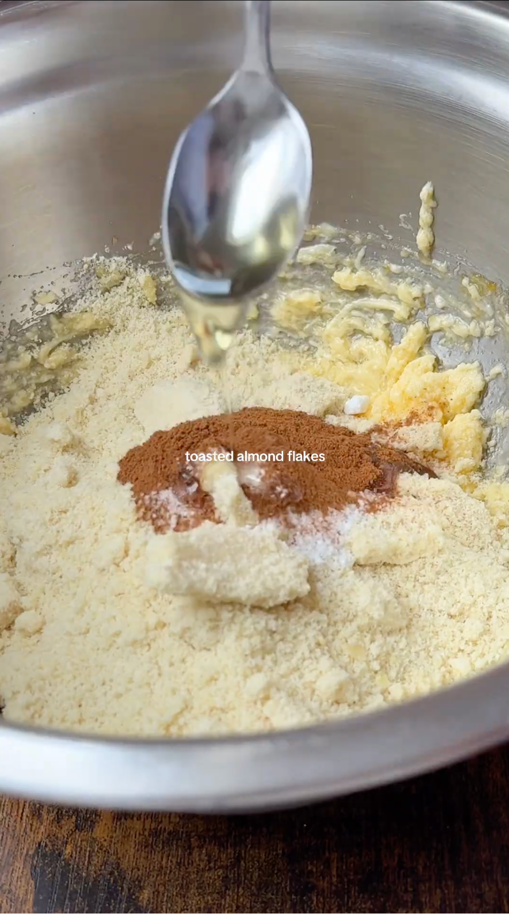 A spoon drizzles liquid into a mixing bowl containing flour, cinnamon, and creamed ingredients. The text toasted almond flakes appears above the mixture.