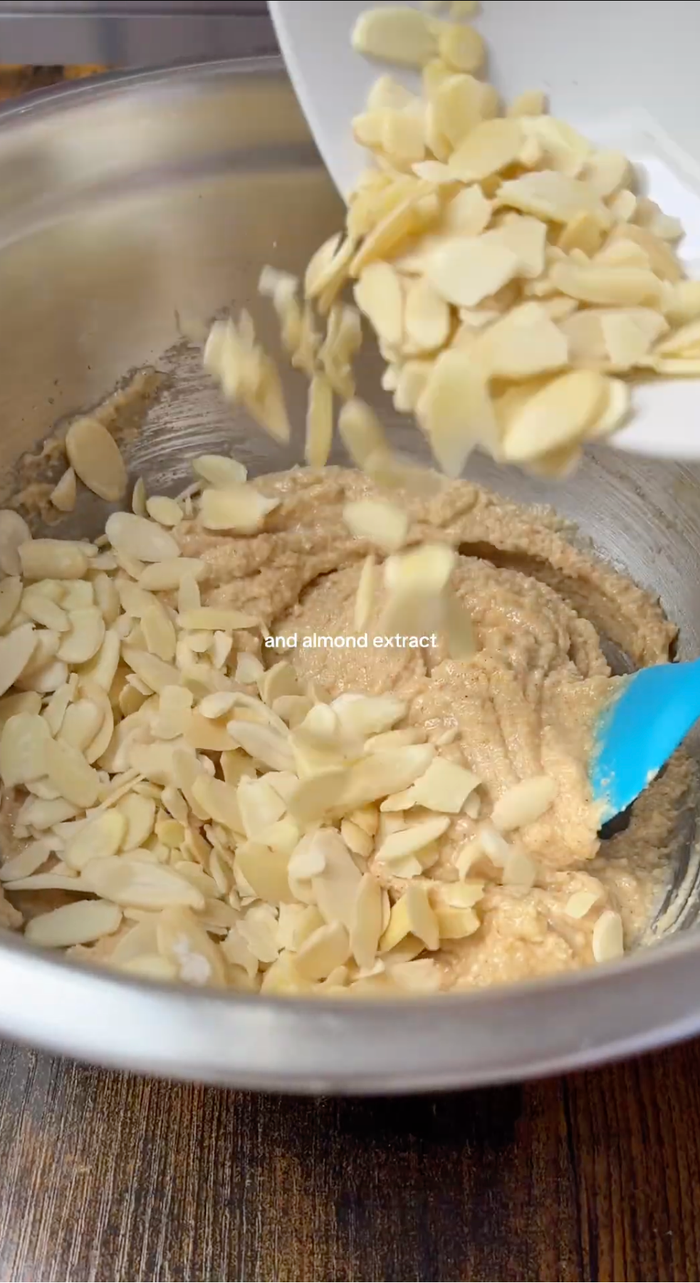 A hand pours sliced almonds into a metal mixing bowl filled with thick, beige batter, using a blue spatula to stir.