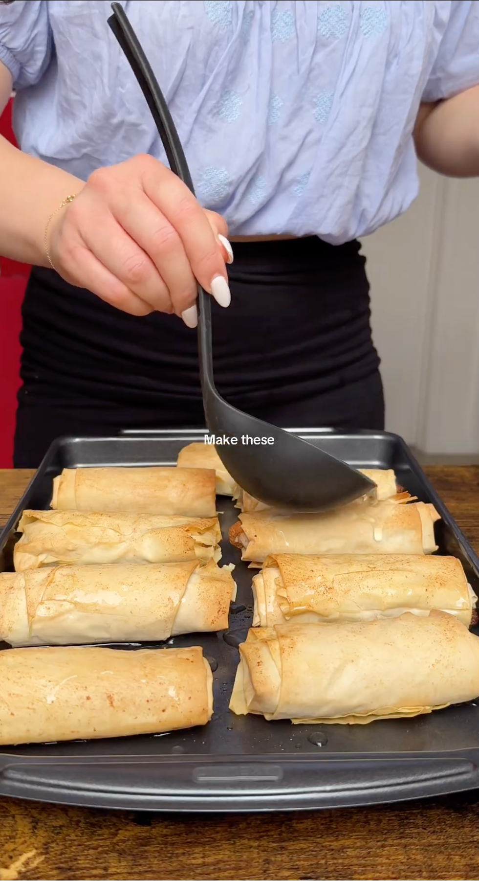A person uses a black ladle to pour liquid over golden-brown rolls on a baking tray. The person is wearing a light blue top and black skirt. Text on the image reads, Make these..