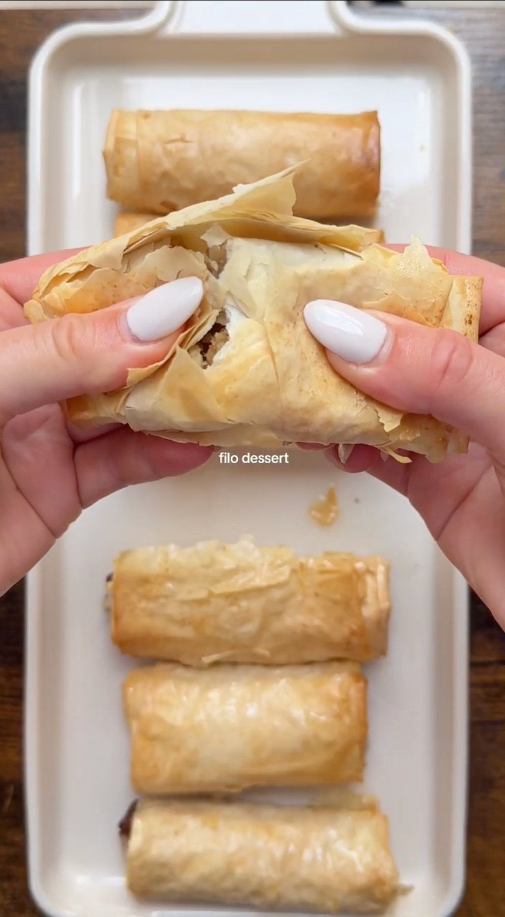 Hands breaking a crispy filo pastry roll in half over a tray with several whole filo dessert rolls placed in a row.