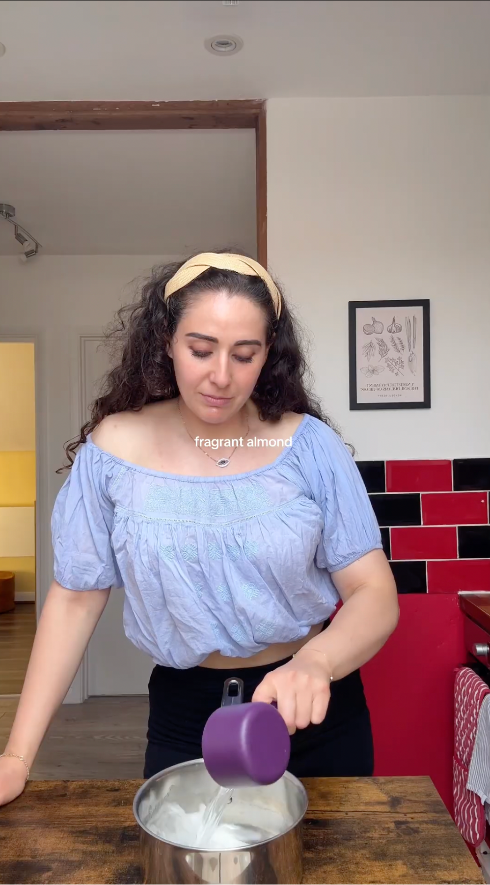 A woman in a blue off-shoulder top pours a white ingredient from a purple measuring cup into a pot on a wooden counter in a kitchen. Text on the image reads fragrant almond..