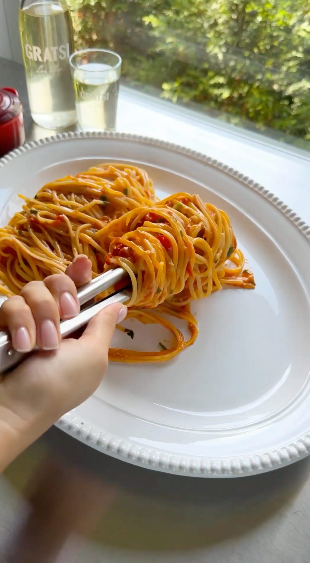 A hand uses tongs to plate a serving of spaghetti with tomato sauce on a white dish near a window with greenery outside.