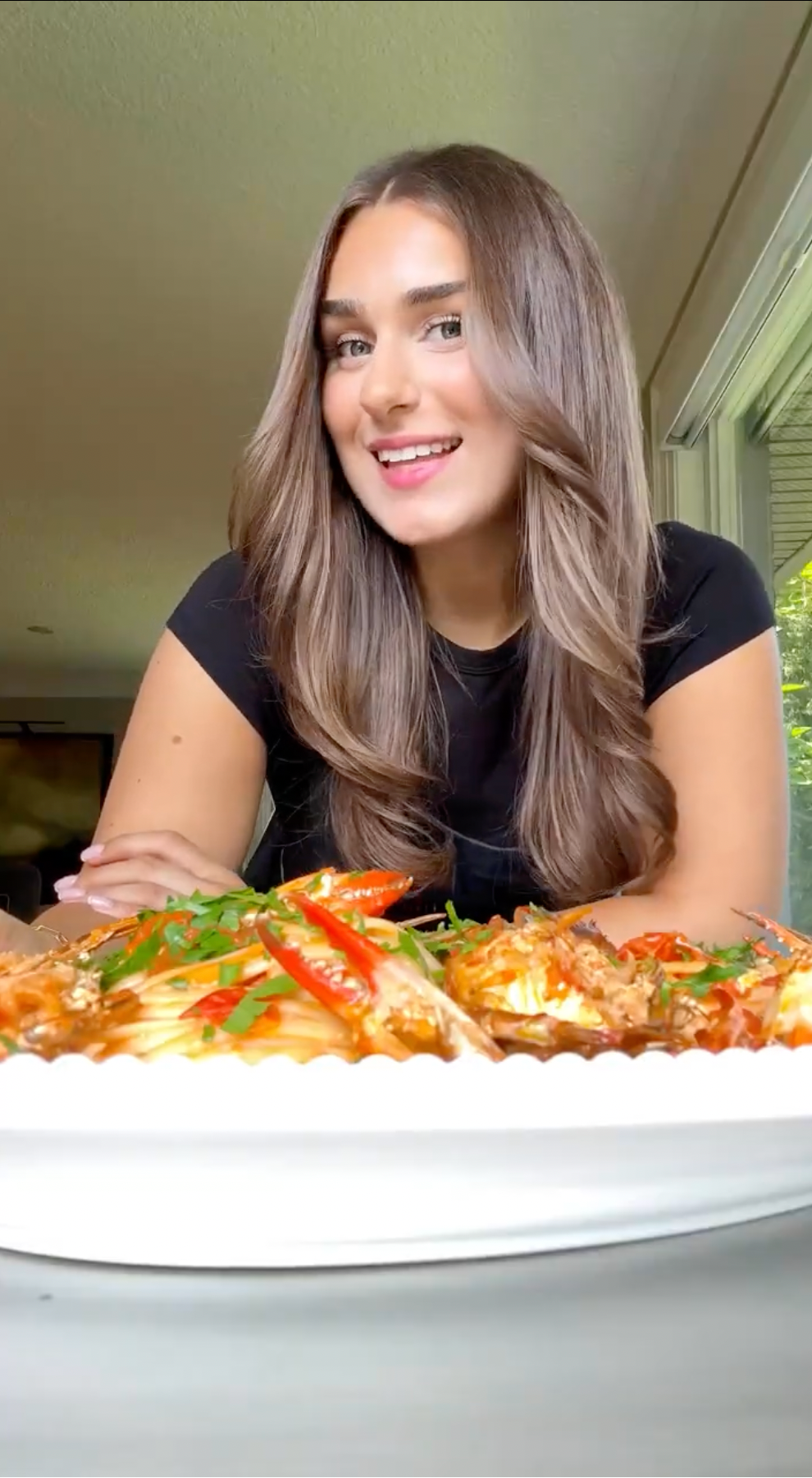 A woman with long brown hair smiles at the camera behind a plate of food topped with sauce, herbs, and sliced chili peppers. She is indoors with natural light coming from a window.