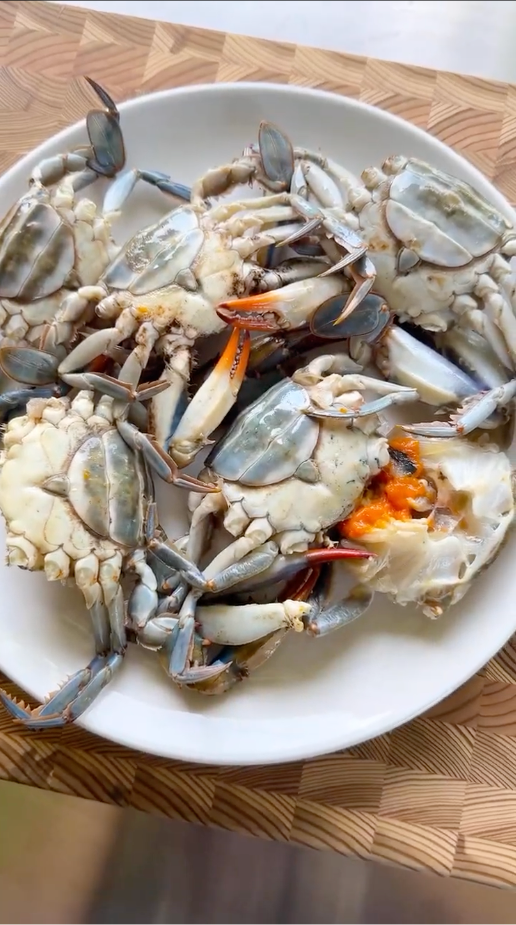 A white plate holds several raw blue crabs, some with orange roe visible, placed on a wooden surface.