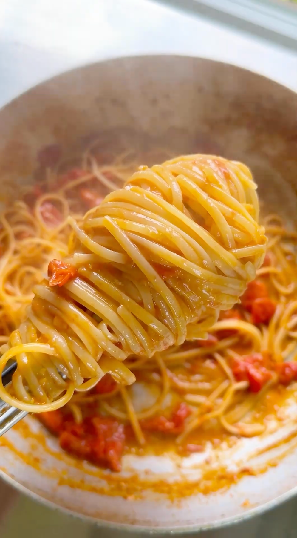 A fork lifts a swirl of spaghetti coated in tomato sauce above a pan filled with more spaghetti and chunks of tomato.