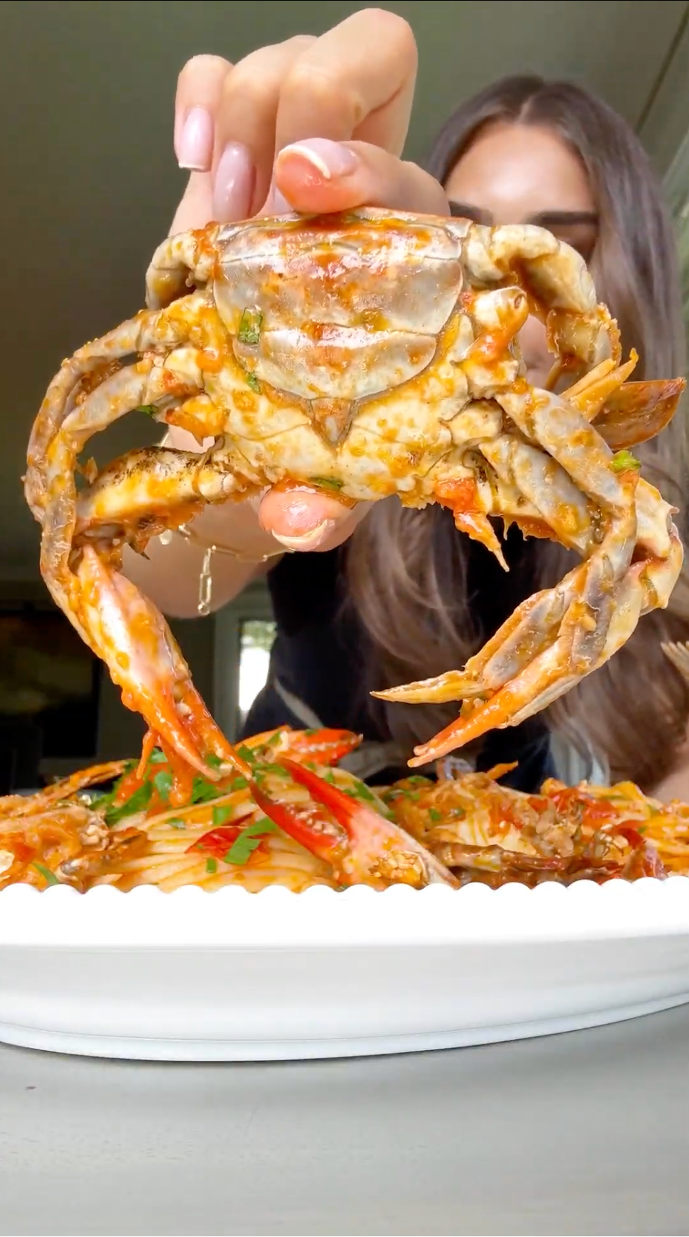 A person holds up a seasoned crab above a plate of saucy seafood garnished with chopped green onions and red peppers.