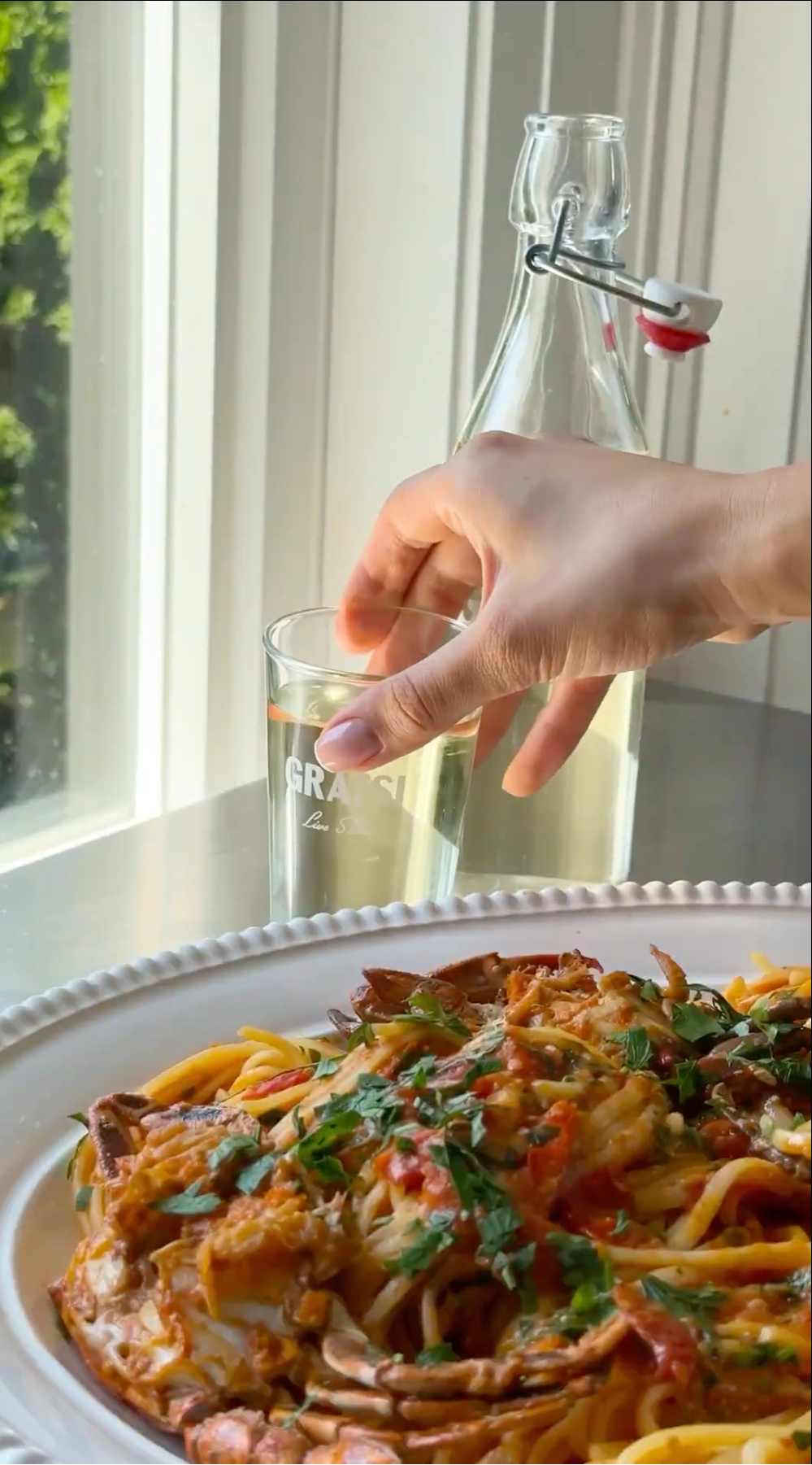 A hand reaches for a glass of water beside a glass bottle on a table near a window, with a plate of pasta topped with herbs and sauce in the foreground.