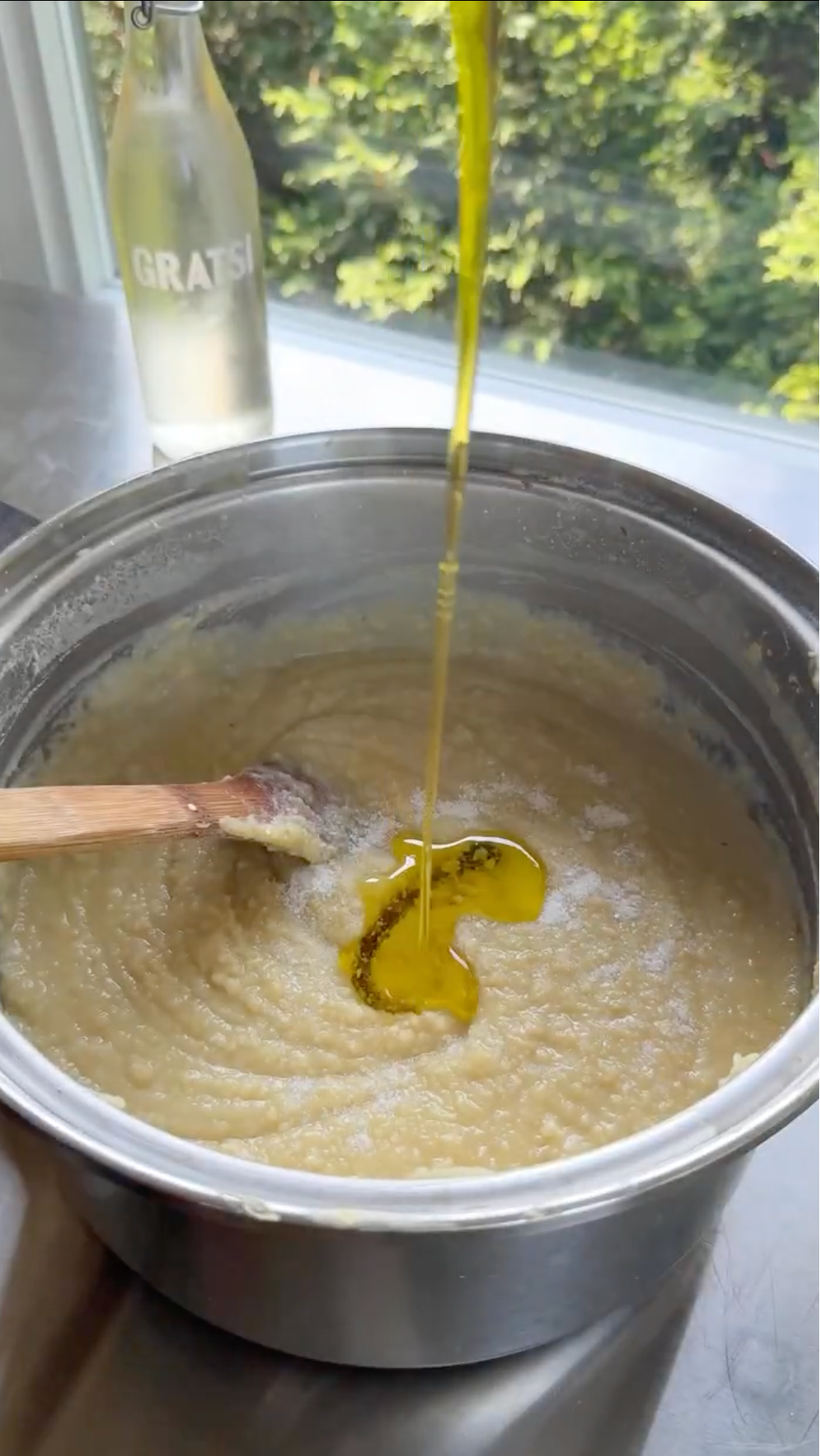 Olive oil is being poured into a pot of creamy mashed potatoes with a wooden spoon inside. A glass bottle is visible in the background near a sunny window with green trees outside.