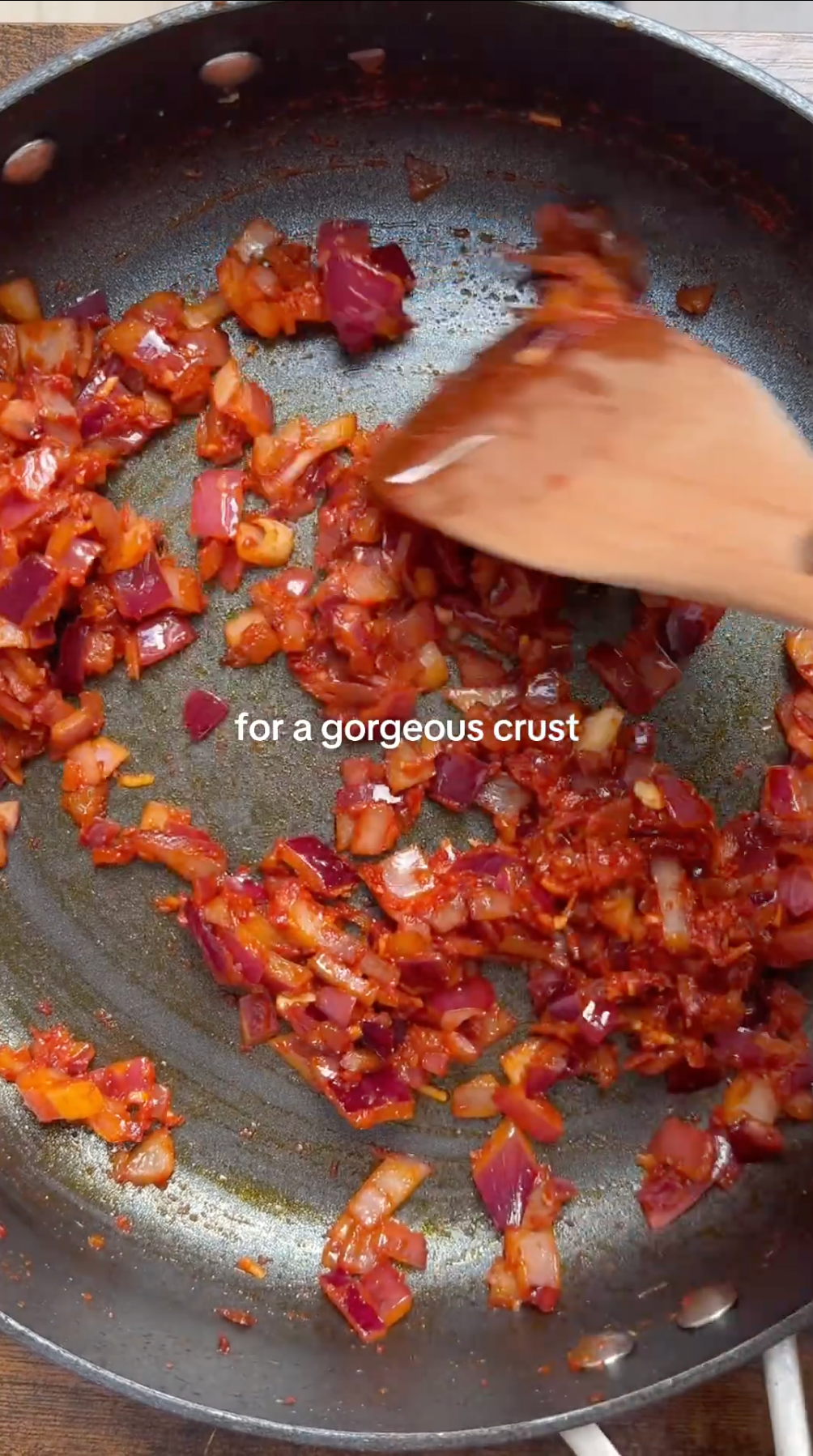 Chopped red onions and tomato paste are being sautéed in a pan with a wooden spatula. The text for a gorgeous crust appears over the mixture.