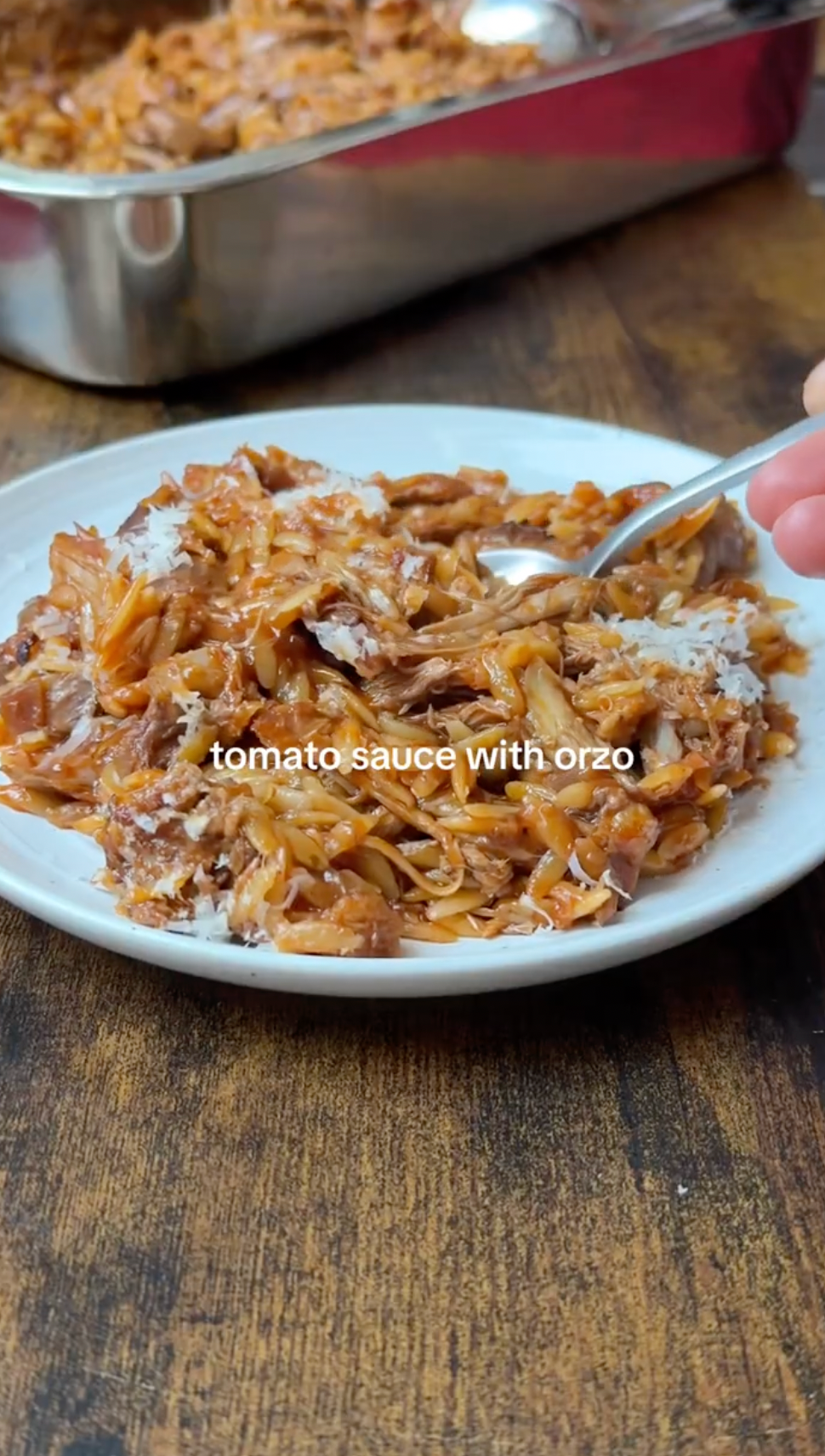 A white plate filled with orzo pasta mixed in tomato sauce, topped with grated cheese. A hand is holding a spoon, ready to take a bite. There is a metal baking dish with more orzo in the background on a wooden table.