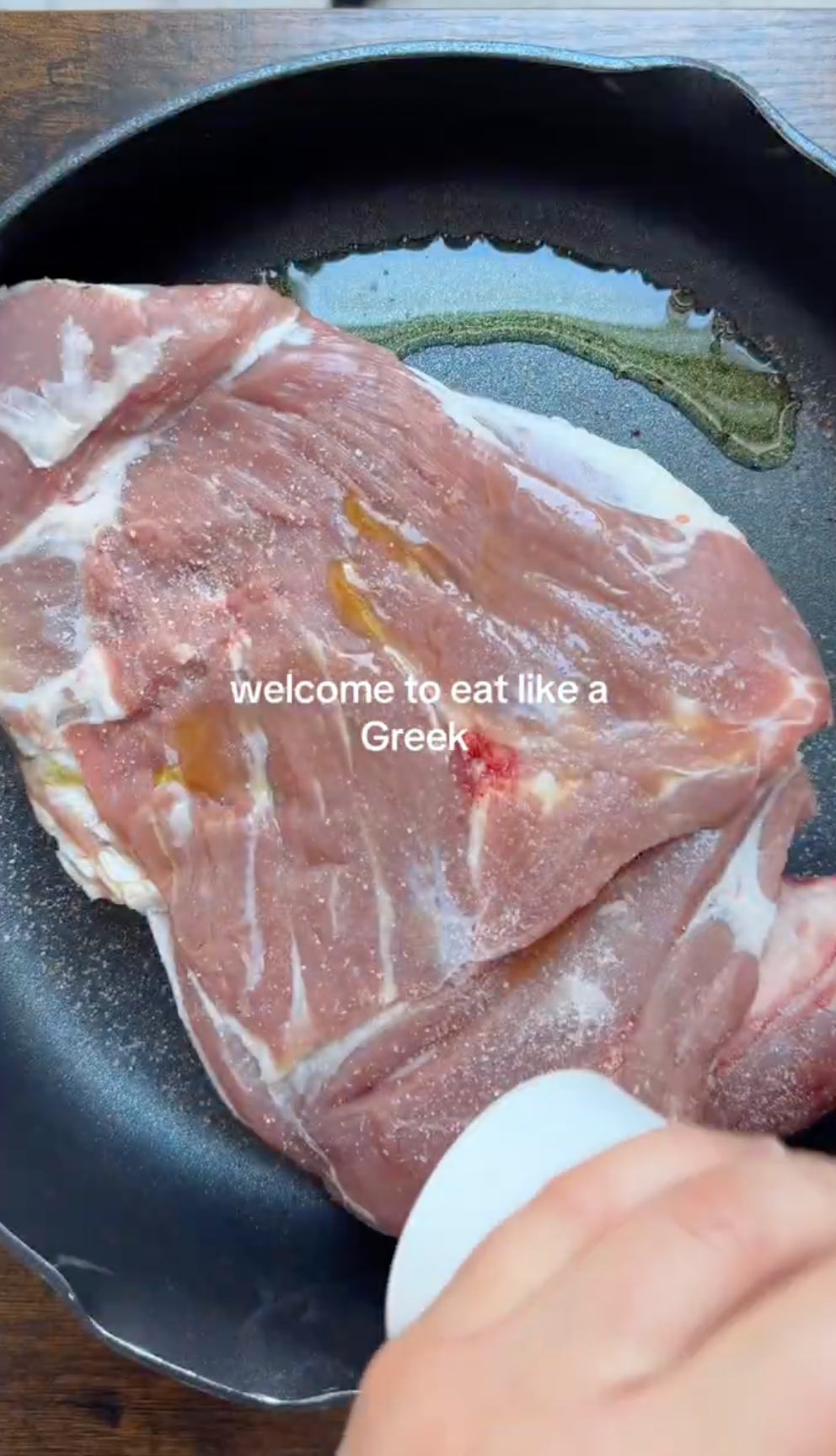 Raw meat is placed in a black skillet with oil. A hand holds a white cup near the meat. The text on the image reads, welcome to eat like a Greek..