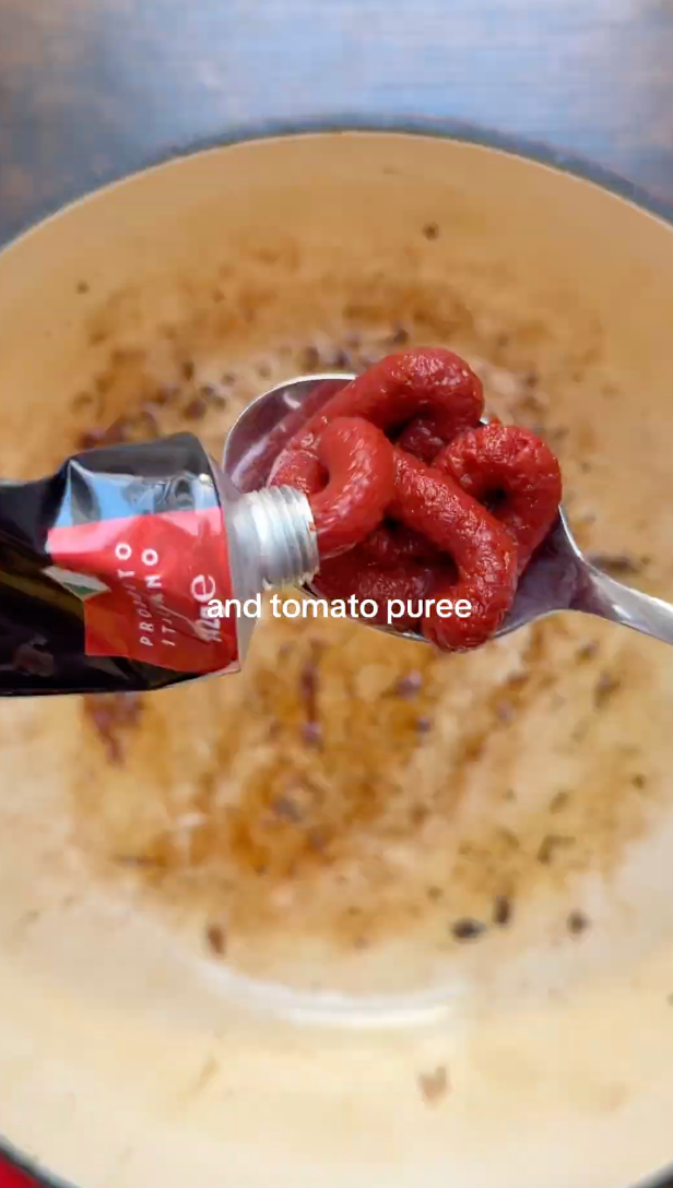 A spoon holds tomato puree squeezed from a tube over a pot with browned bits at the bottom. Text on the image reads, and tomato puree..