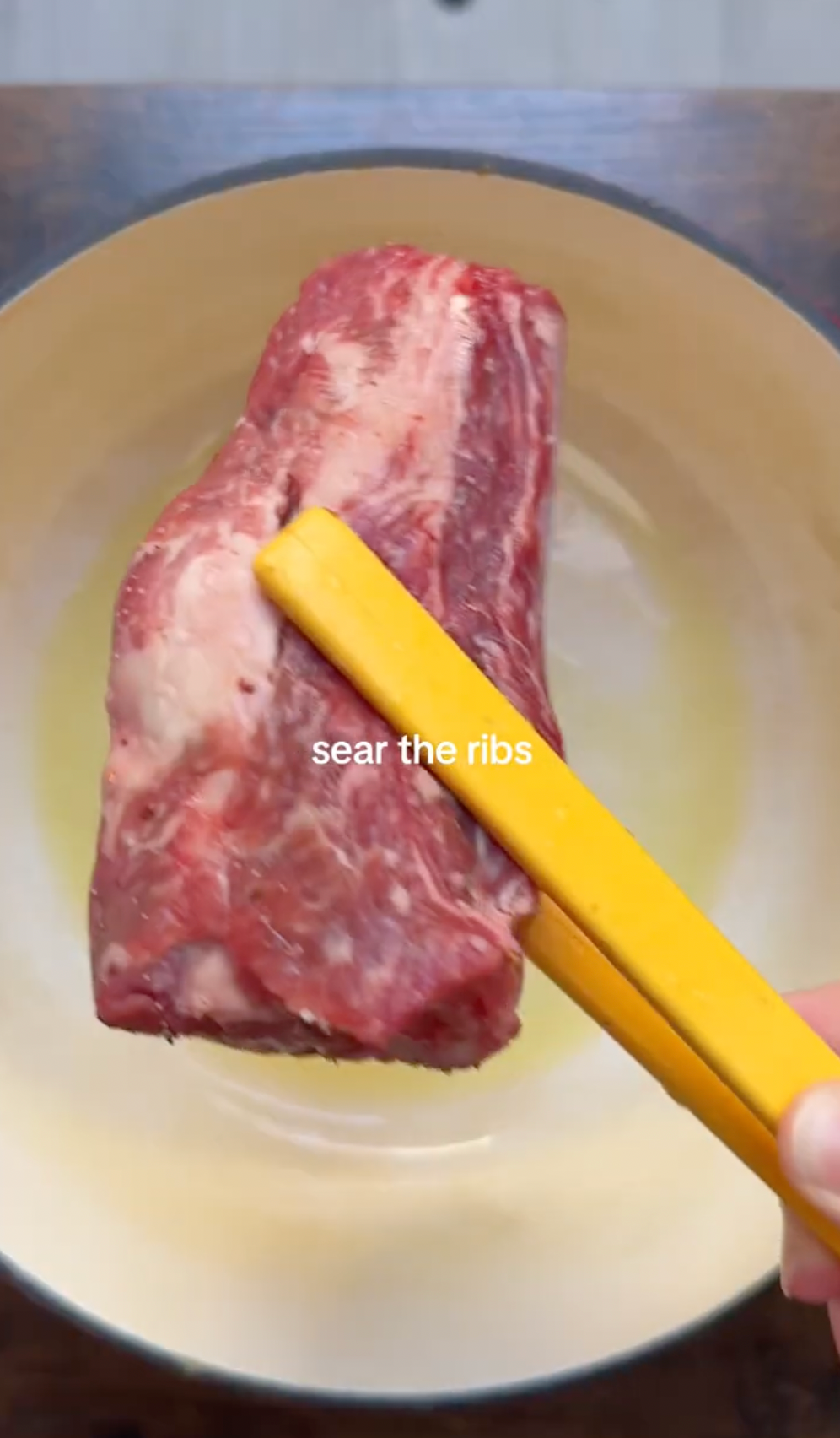 A hand holding raw beef ribs with yellow tongs over a light-colored pan, about to sear the meat. The text sear the ribs is displayed in the center of the image.