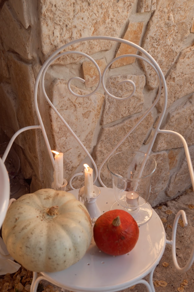 A white metal chair holds two lit candles, a clear glass candle holder with a tea light, a white pumpkin, and a small orange pumpkin.
