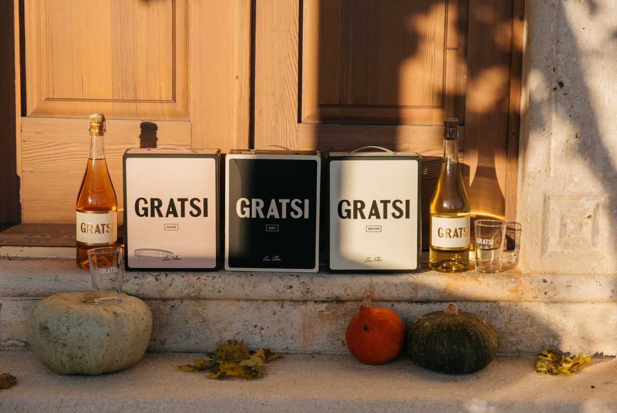 Two bottles of wine, three boxed wine containers labeled GRATSI, and two glasses arranged on stone steps with three pumpkins.