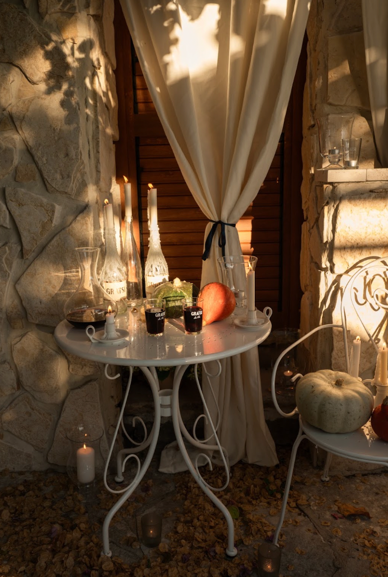 A white bistro table set for two with candles, glasses of red wine, and a pumpkin, placed on a patio with a stone wall and curtains.