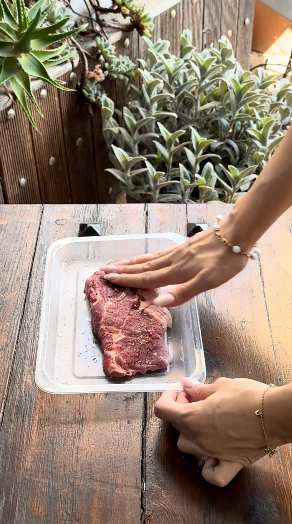 A person seasons a raw steak in a plastic tray on a wooden table.
